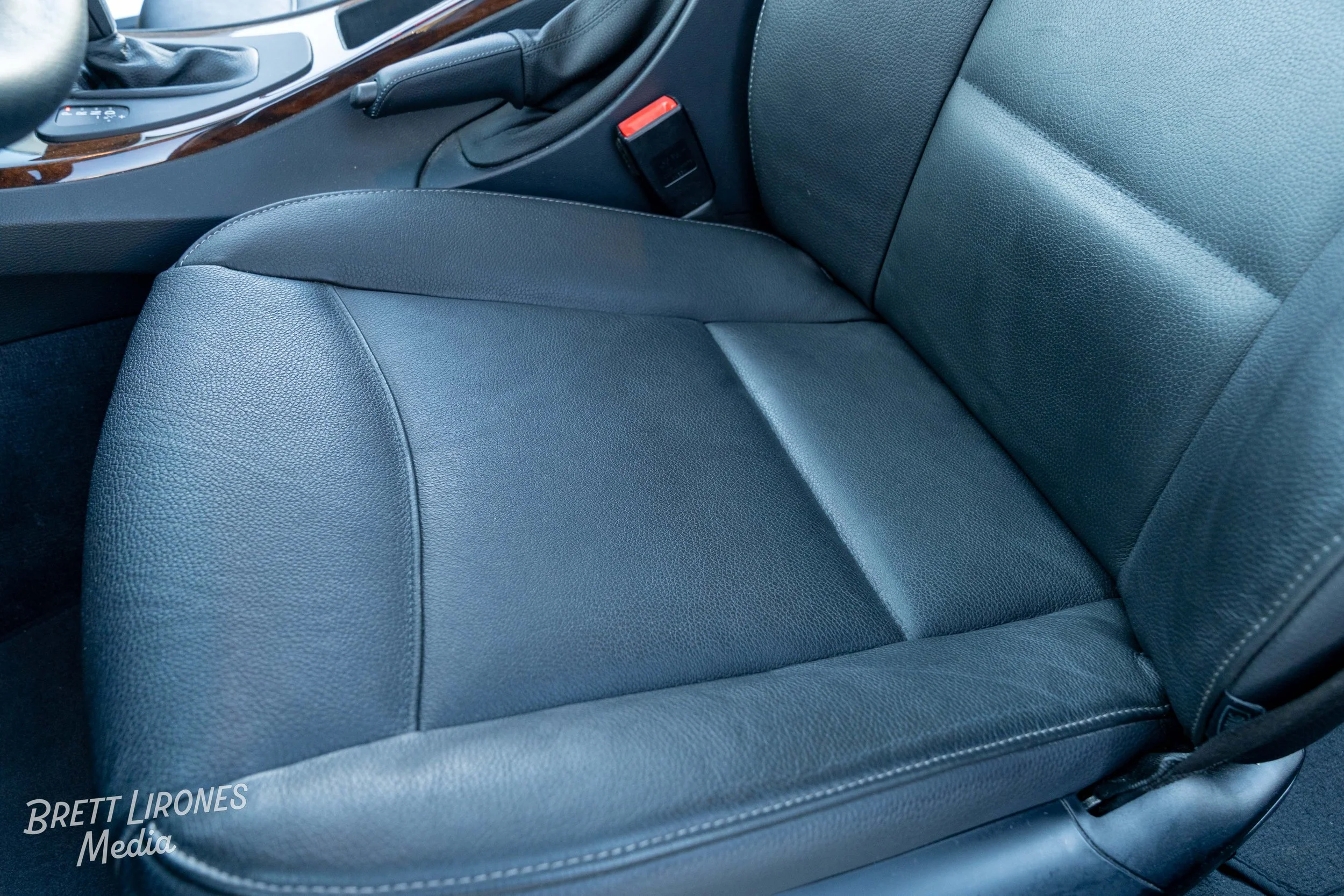 Close-up of a black leather car seat with textured upholstery and stitching visible, including part of the center console with a gear shift and seatbelt buckle.