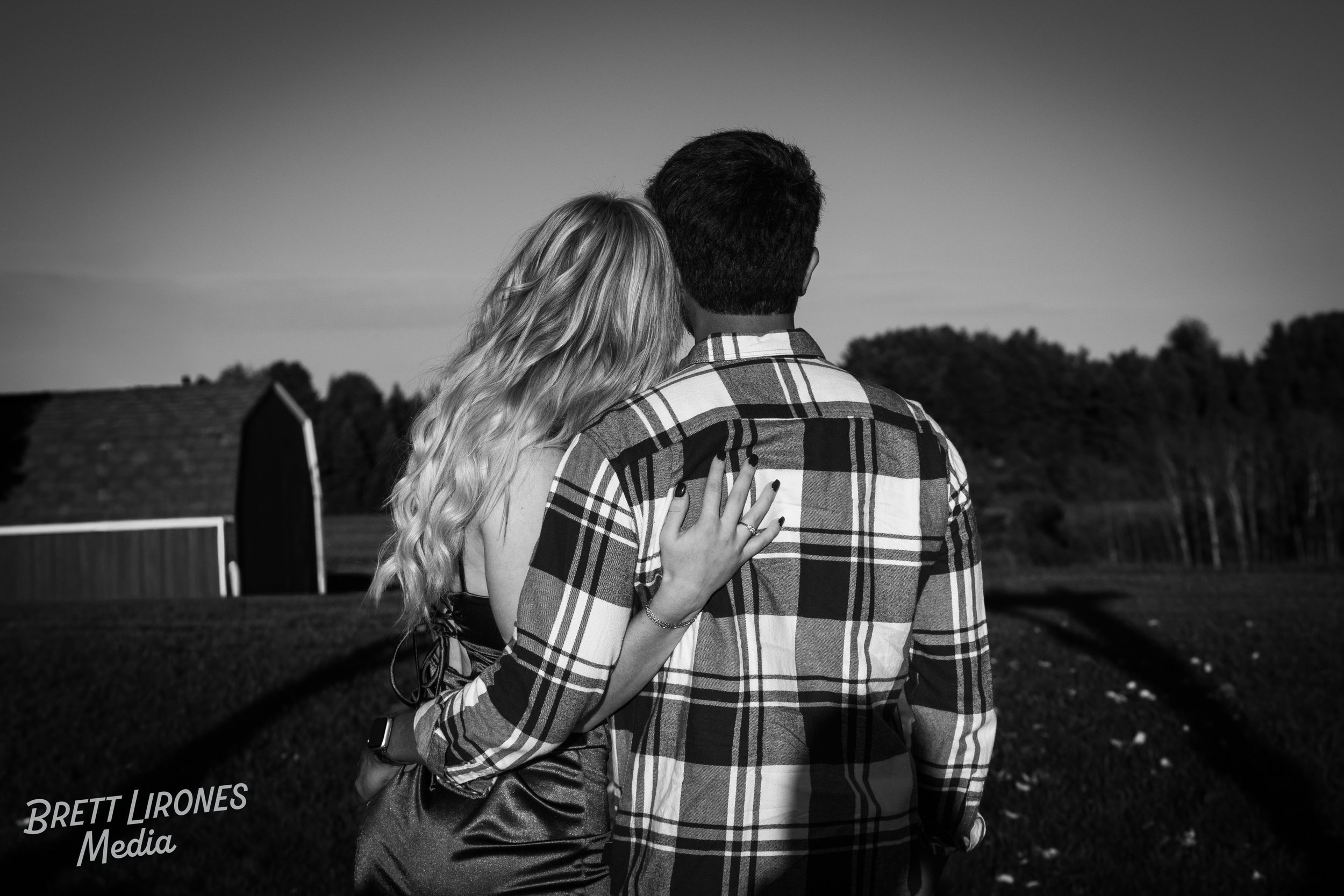 A black and white photo of a couple standing outdoors, facing away from the camera. The woman has long, wavy hair and is wearing a dress, while the man has short hair and is wearing a checkered shirt. They are embracing each other with the woman's ha