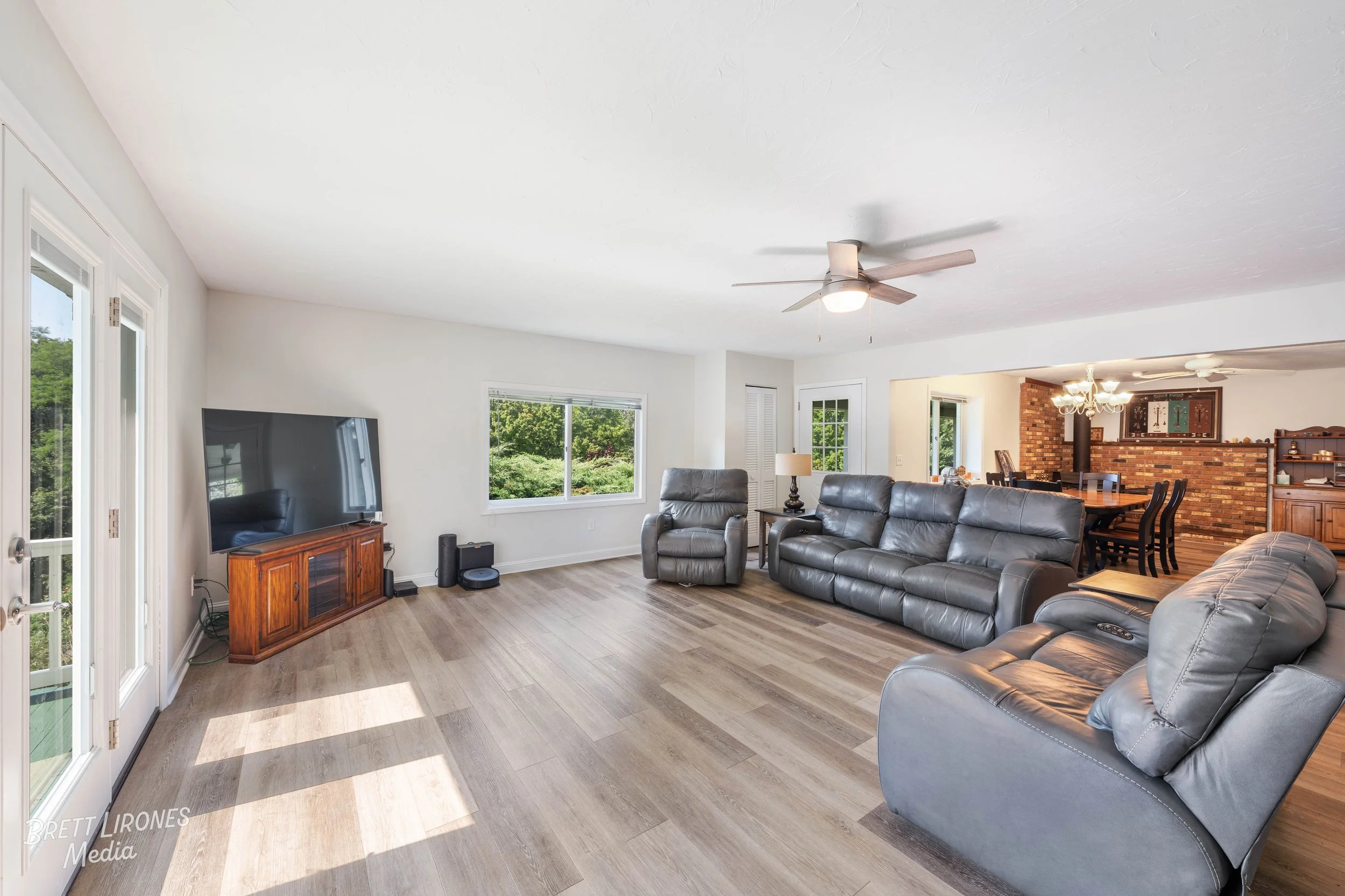 Living room with gray leather furniture, a flat-screen TV on a wooden stand, large windows with greenery outside, laminate flooring, ceiling fan, and a view into the dining area with a brick accent wall and chandelier.