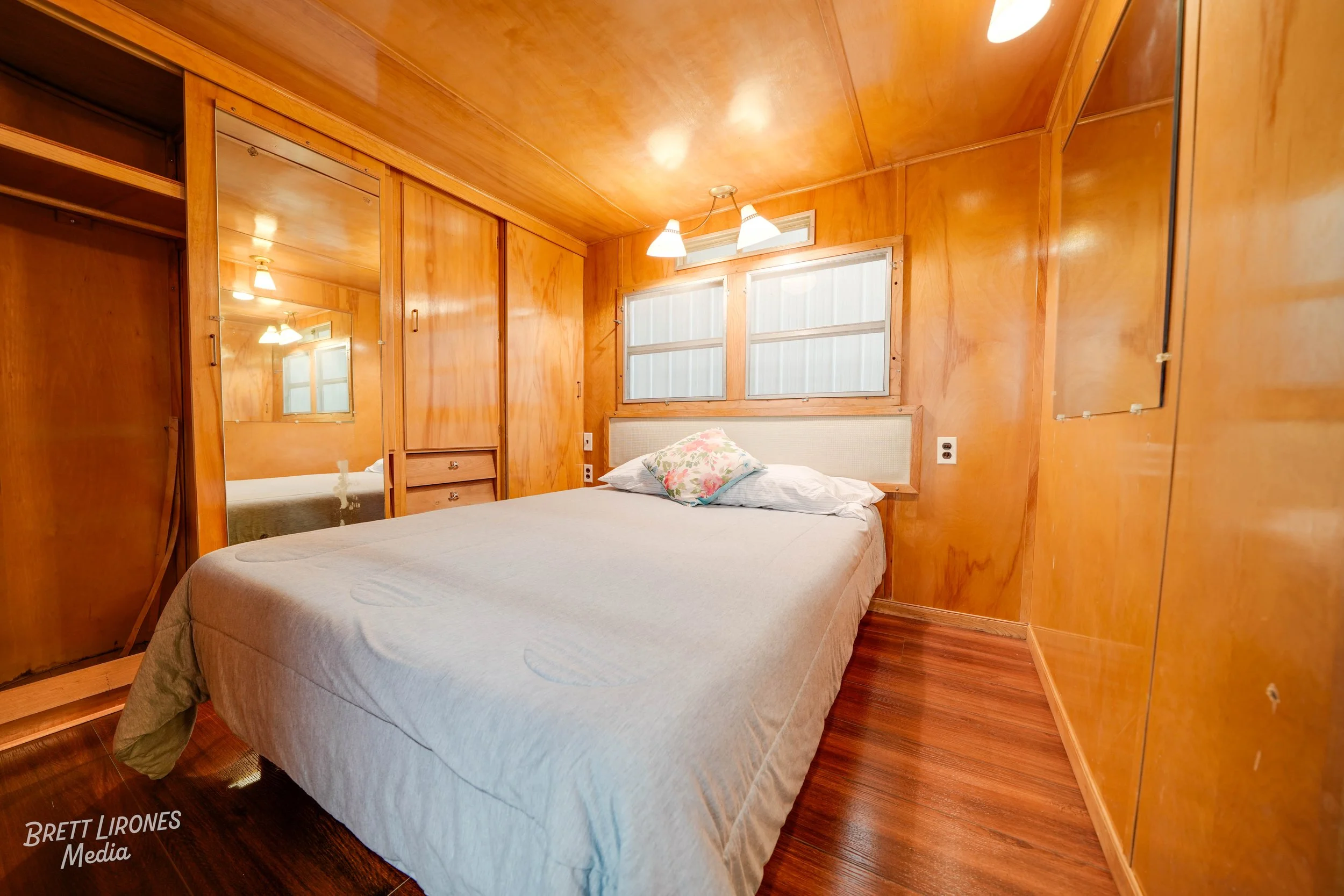 A small bedroom with wood-paneled walls and ceiling, a window with blinds, a white headboard, and a bed with white bedding and floral pillows.