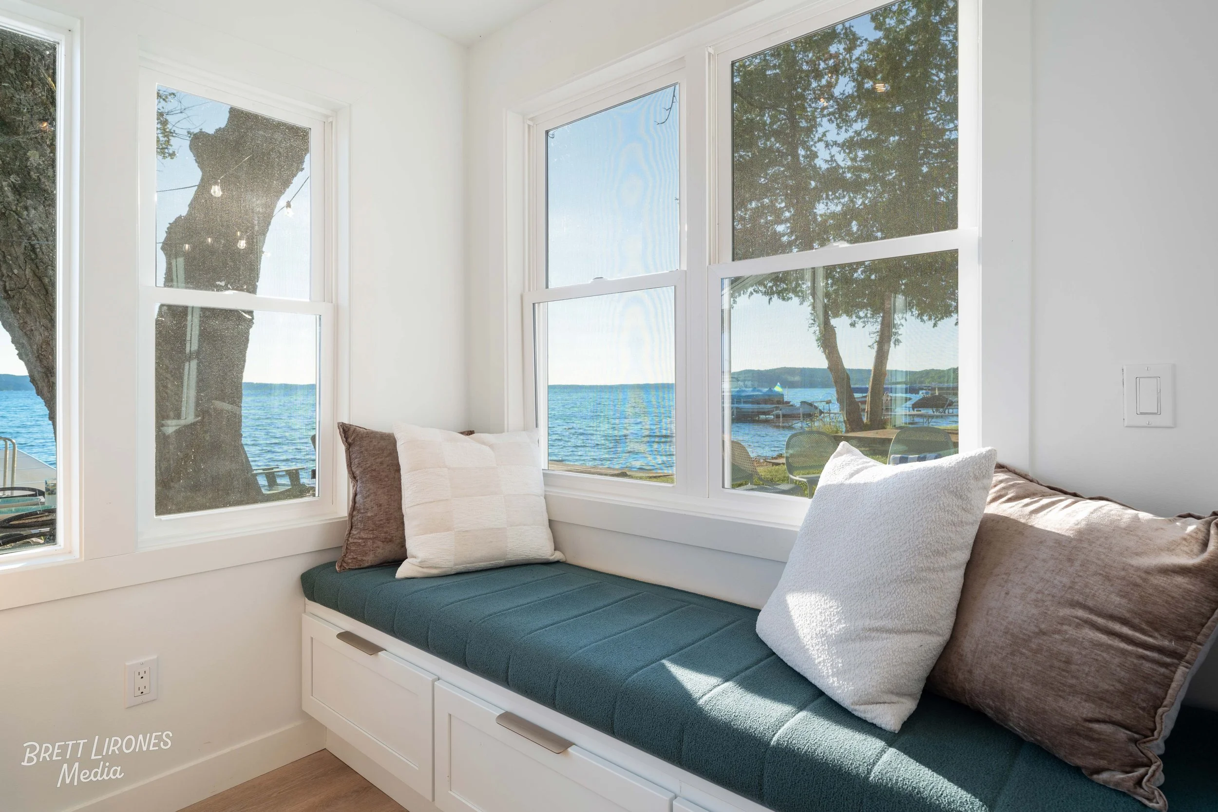 Indoor window seat with pillows facing a water view through large windows, showing trees, boat docks, and water