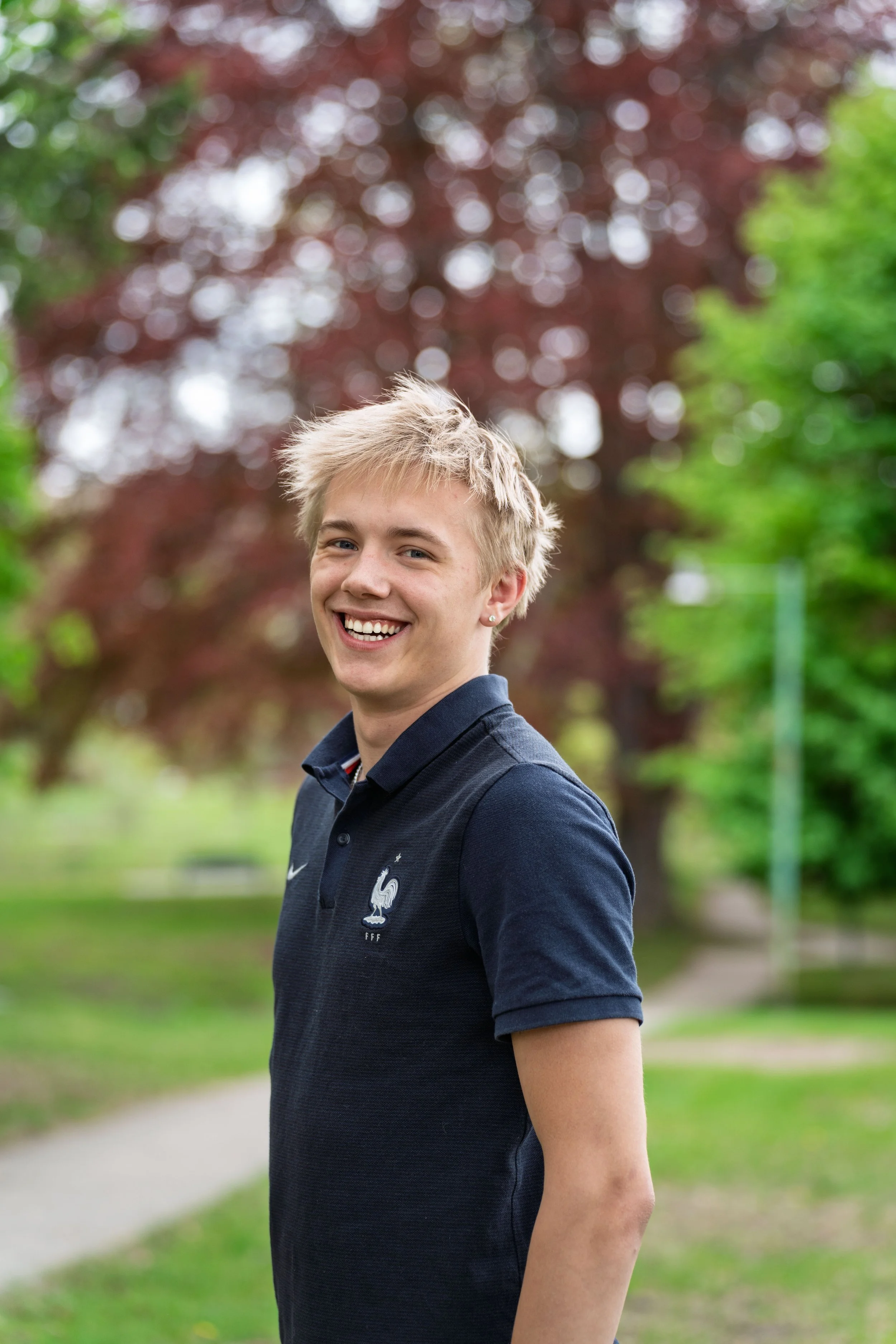 Young man smiling outdoors with green trees and a pathway in the background.