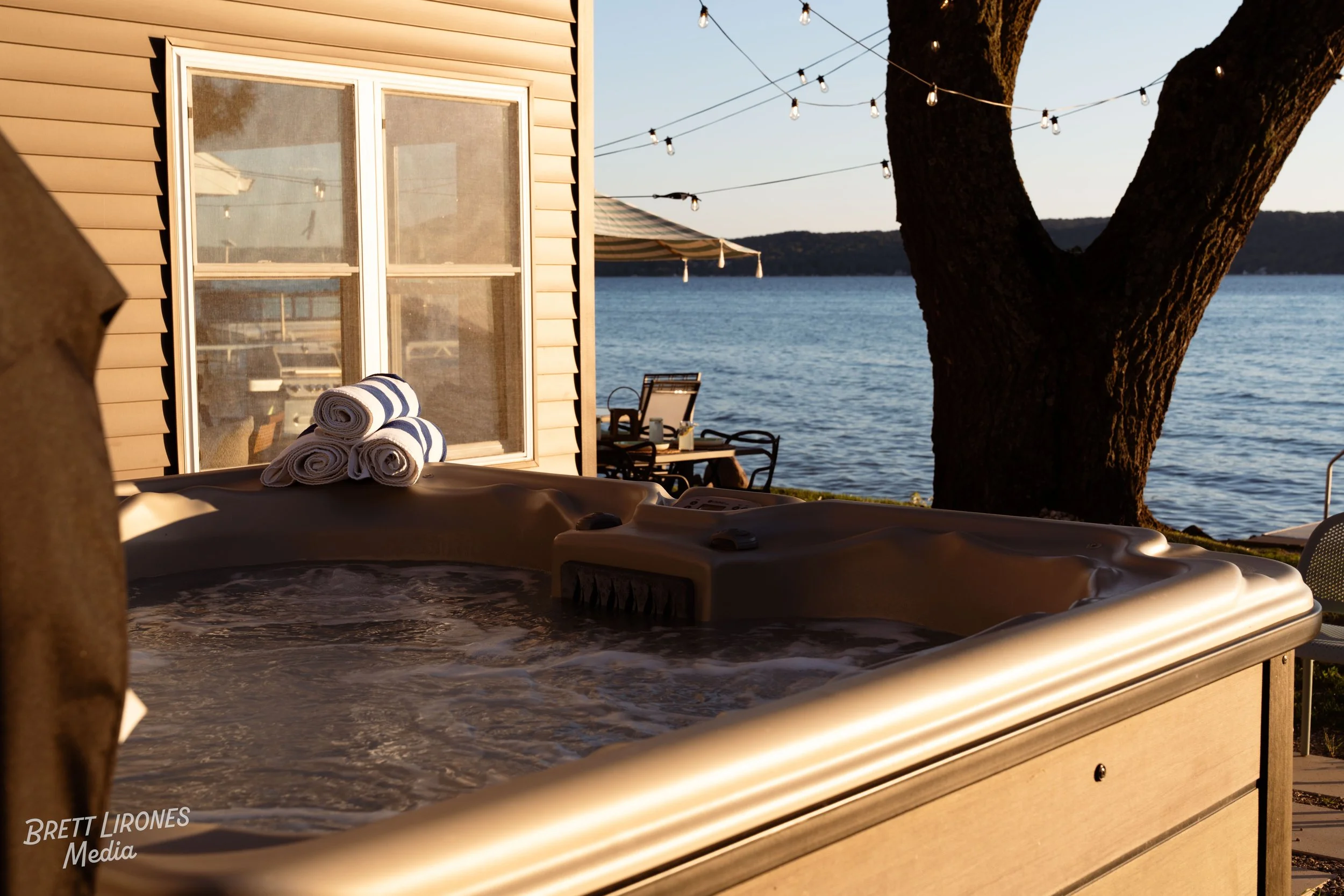An outdoor hot tub on a wooden deck by a lake at sunset with a large tree and string lights overhead, towels rolled on the hot tub, and a house with a window in the background.