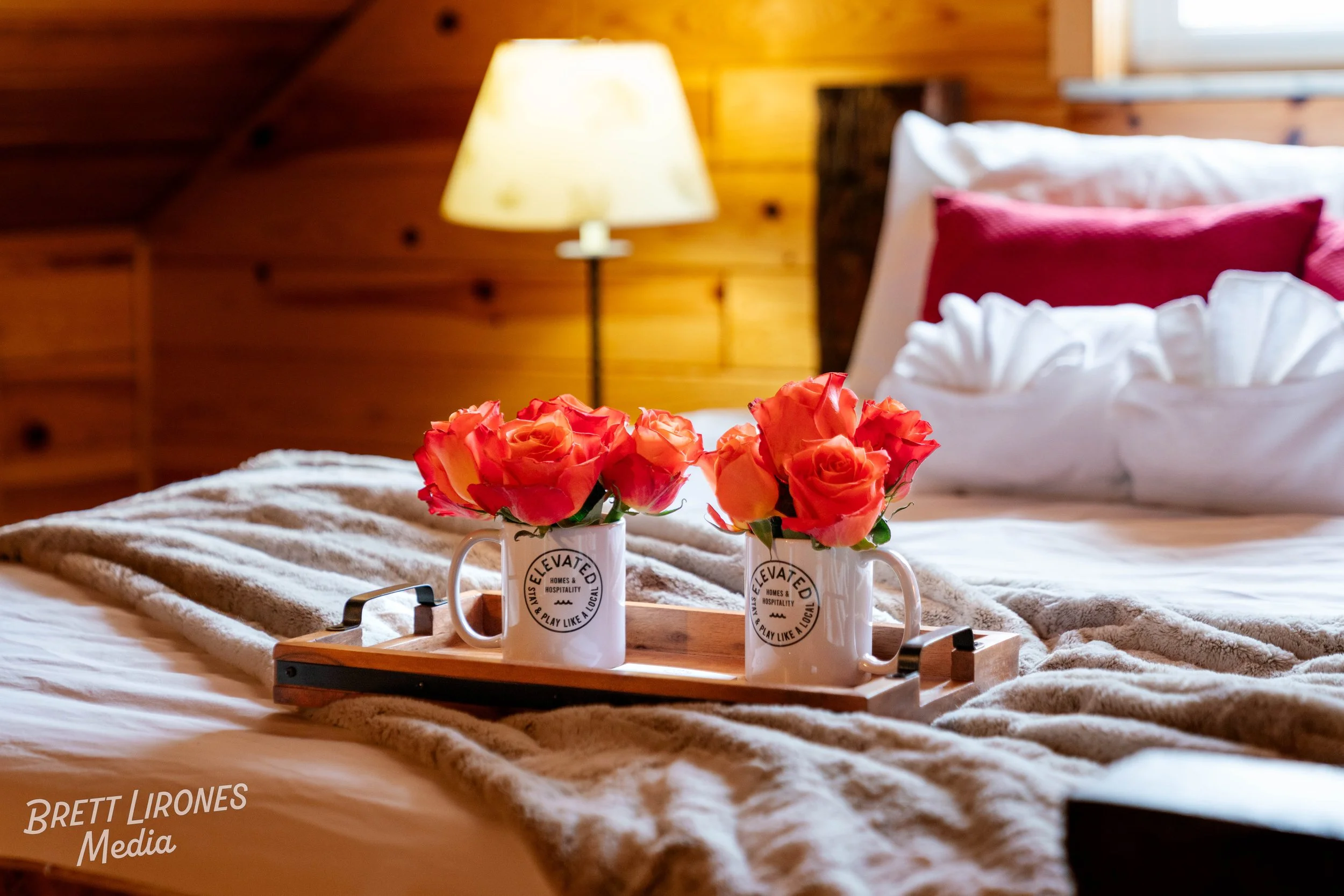 A cozy bedroom with a bed, pillows, and a tray with two mugs of pink roses, wooden walls, a bedside lamp, and a window.
