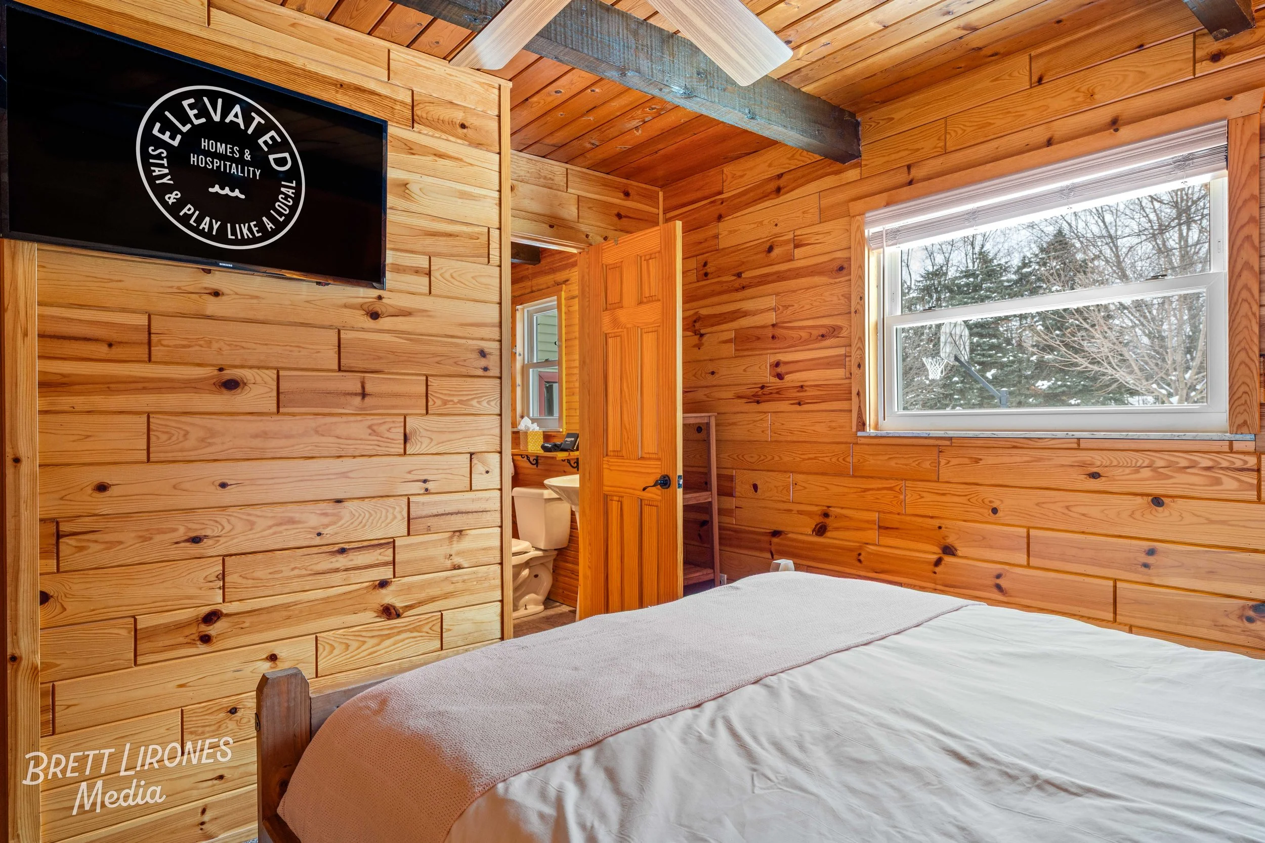 A cozy bedroom with wooden panel walls and ceiling, a bed with white sheets and a light pink blanket, a window with a view of trees and a basketball hoop outside, and a TV mounted on the wall displaying a black background with white text.