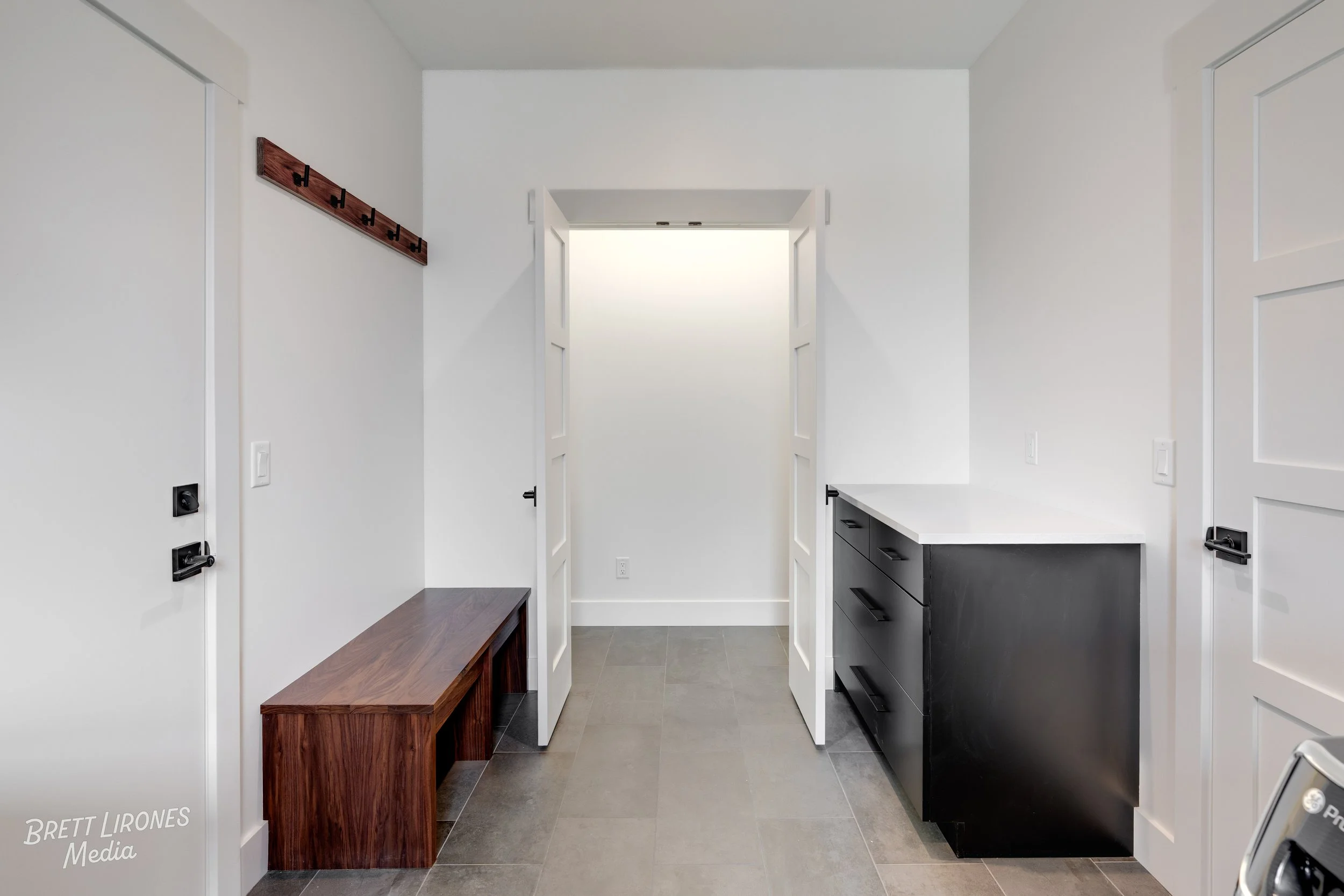 Modern laundry room with white walls, gray tiled floor, white double doors, black cabinet with drawers, and a wooden bench with hooks on the wall.