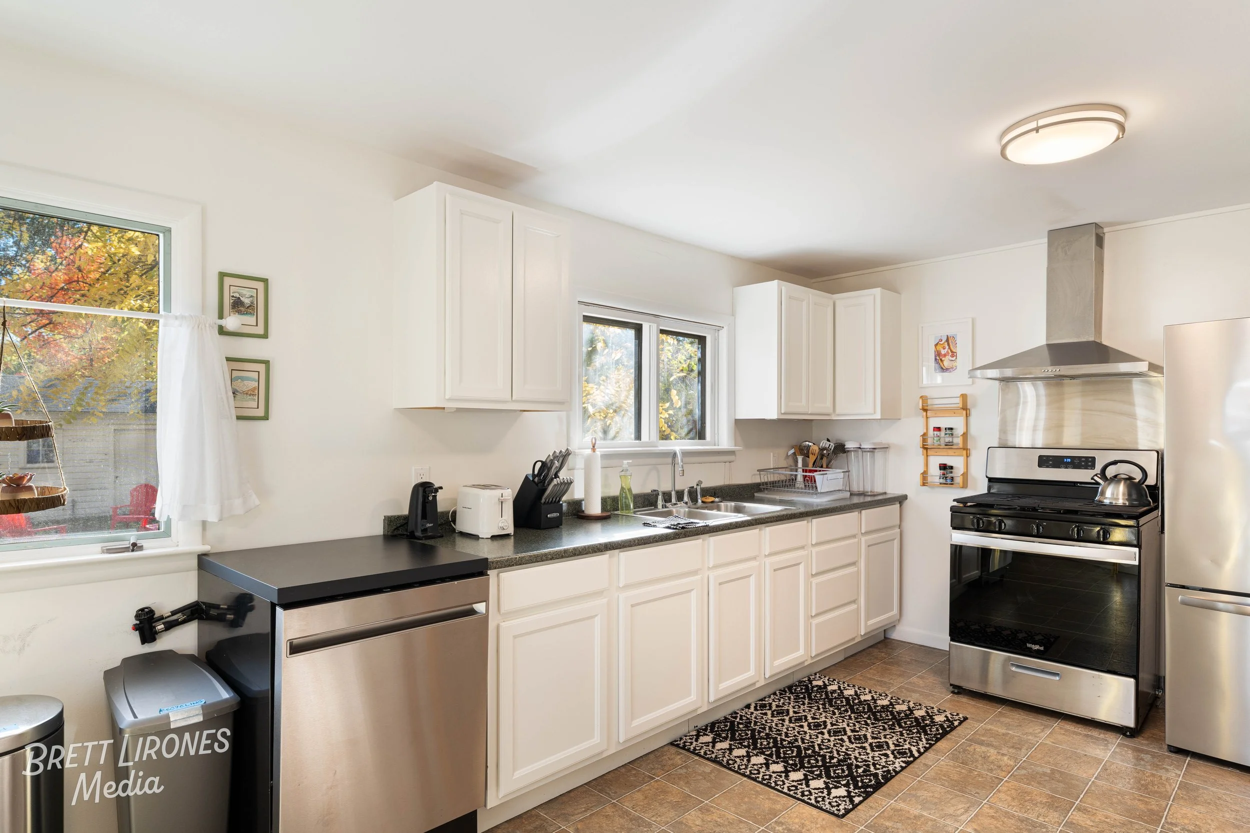 A clean kitchen with white cabinets, black countertops, stainless steel appliances including a stove and refrigerator, and a window with a view of autumn trees.