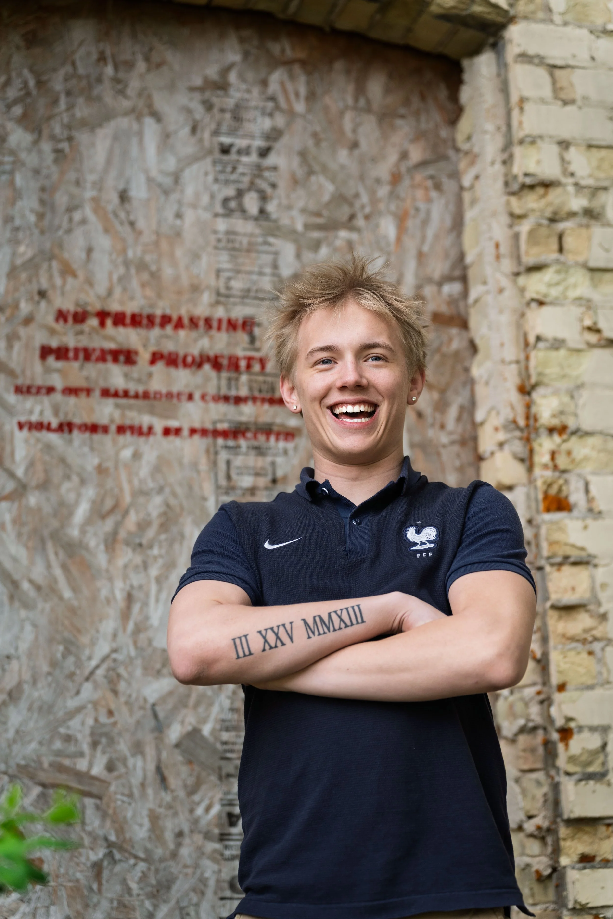 A young woman with short blonde hair smiling with arms crossed, standing outdoors in front of a brick wall with red and black text, wearing a navy blue polo shirt with a rooster emblem.