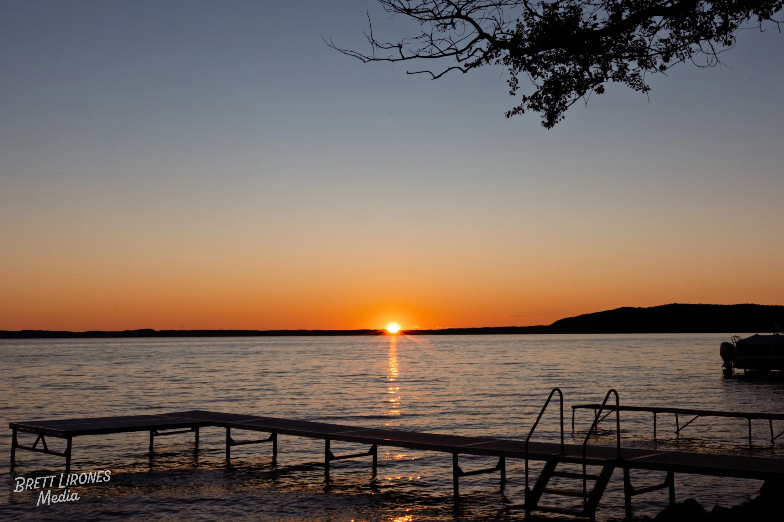 Sunset over a lake with a dock and a boat, silhouette of trees, and hilly landscape in the distance.