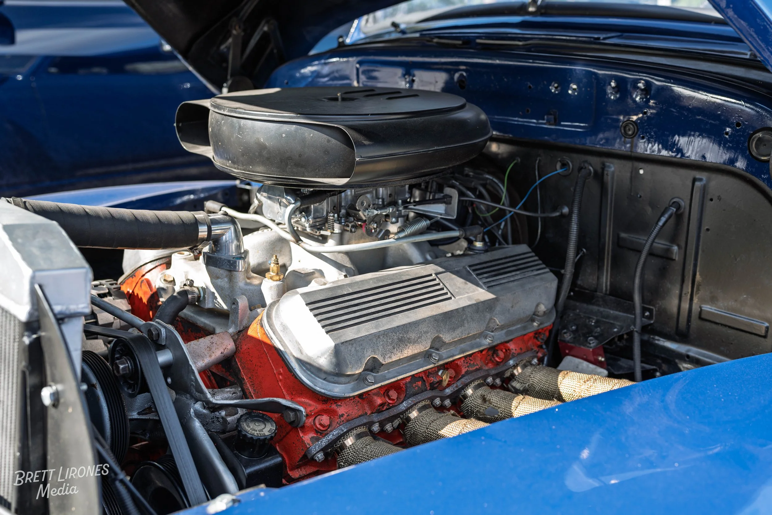 Close-up of a vintage car engine inside the engine bay with a black air filter, metallic engine components, and blue exterior paint.