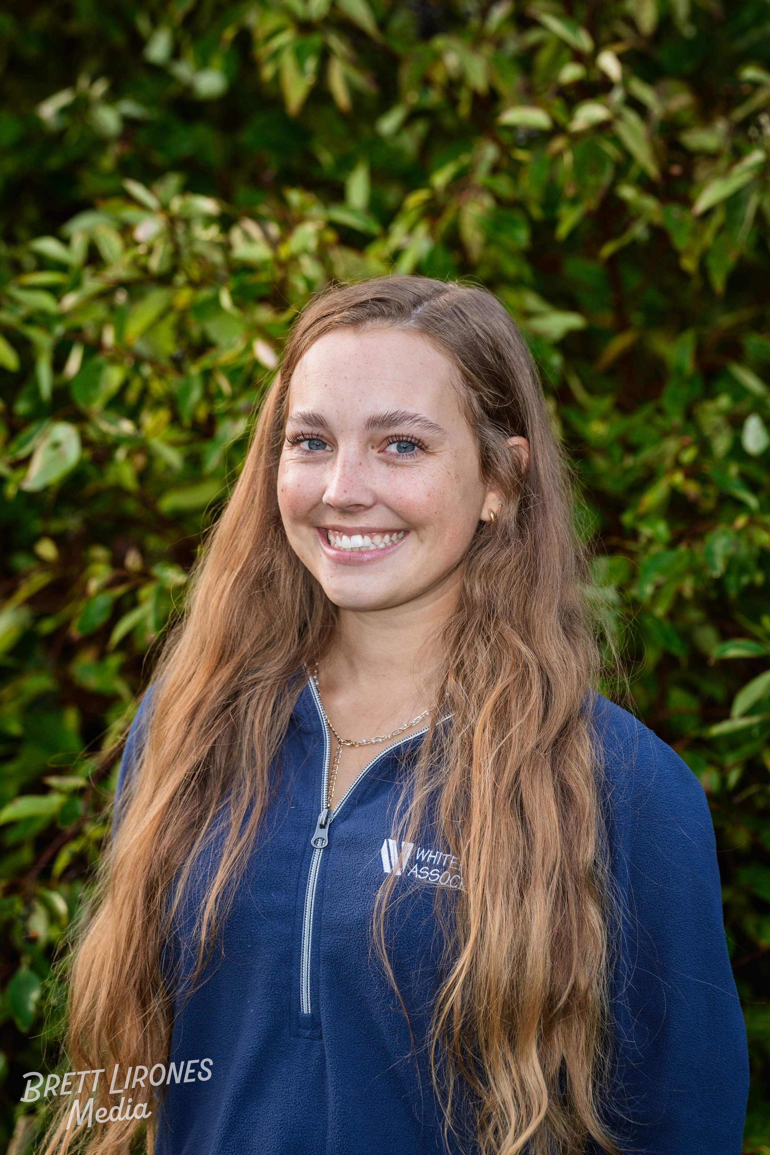 A woman with long, wavy red hair, blue eyes, and fair skin, smiling, wearing a blue zip-up jacket with 'White Pass' logo, standing outdoors in front of green leafy bushes.