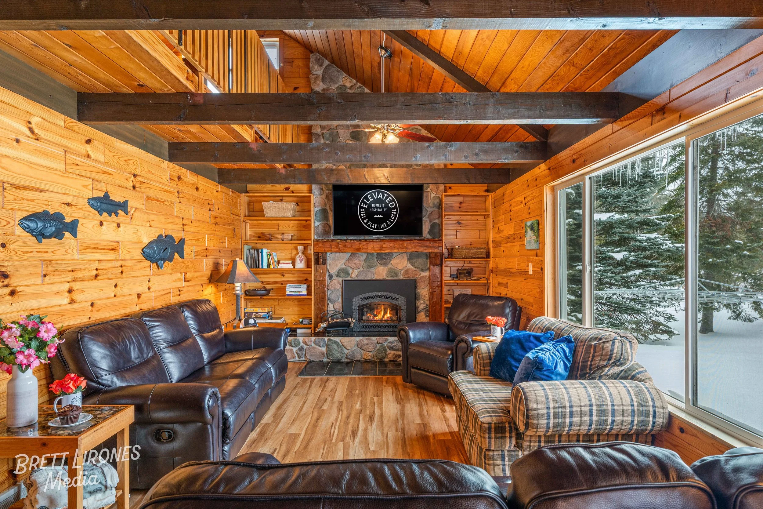 Living room with wooden walls, large windows showing snow-covered trees, leather and plaid furniture, a stone fireplace with a television above, and wall decorations of fish sculptures.