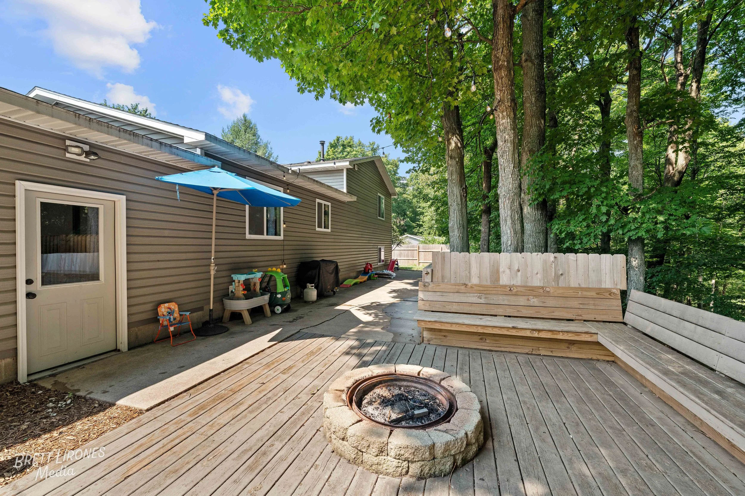 Backyard patio area with a wooden deck, a fire pit, a built-in wooden bench, and a house with gray siding. There is a blue patio umbrella, children's toys, and lush green trees providing shade.