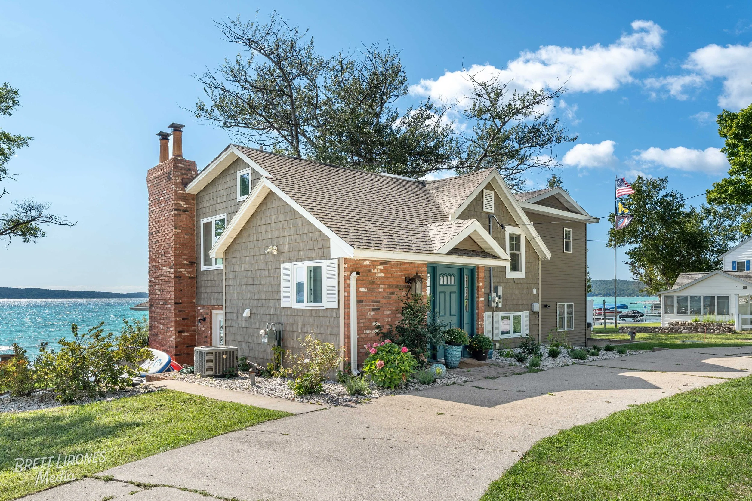 A house with gray shingle siding, brick chimney, and a teal front door, situated near a body of water, with a lawn, potted plants, and a clear blue sky.