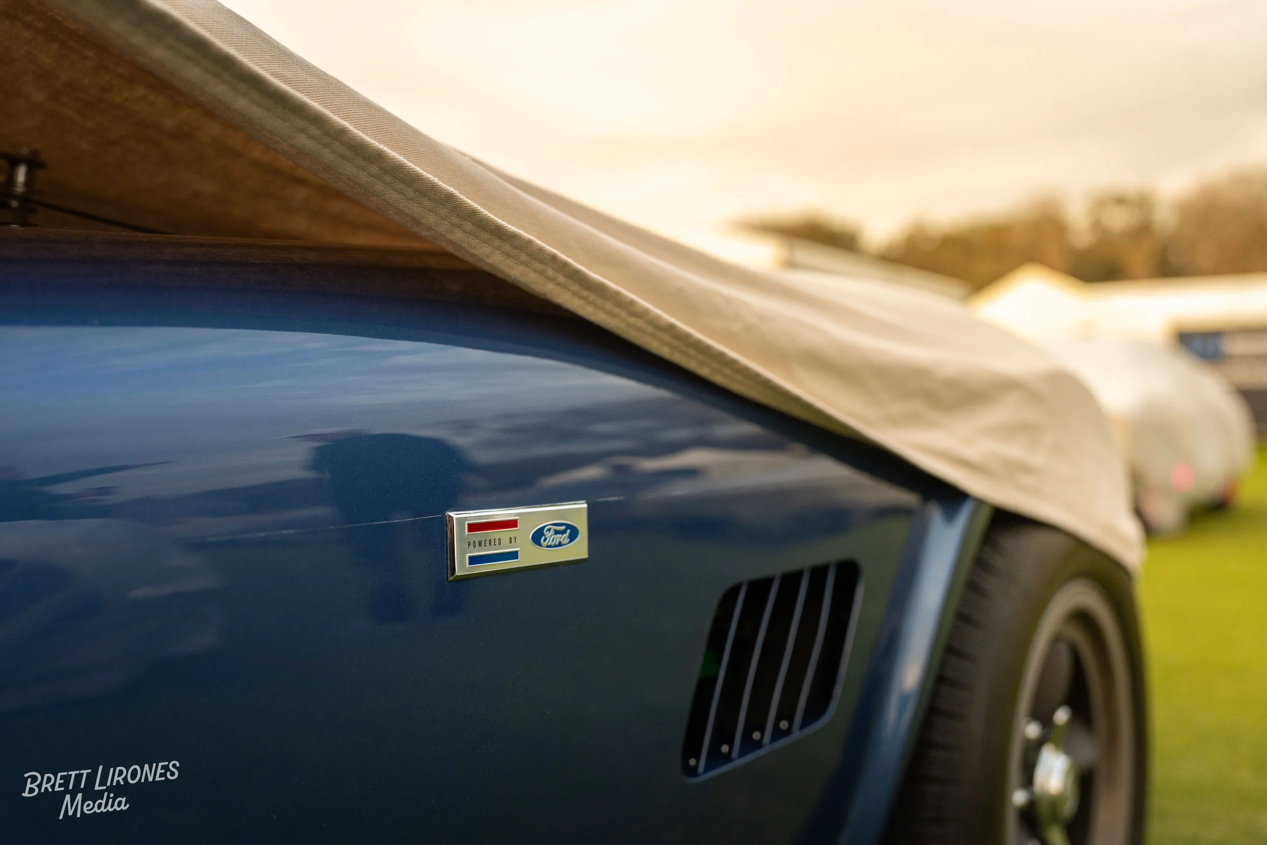 Close-up of a blue vintage vehicle with a Ford emblem and a 'Powered by Ford' badge, partially covered by a beige canvas cover, parked on grass with other vehicles in the background.