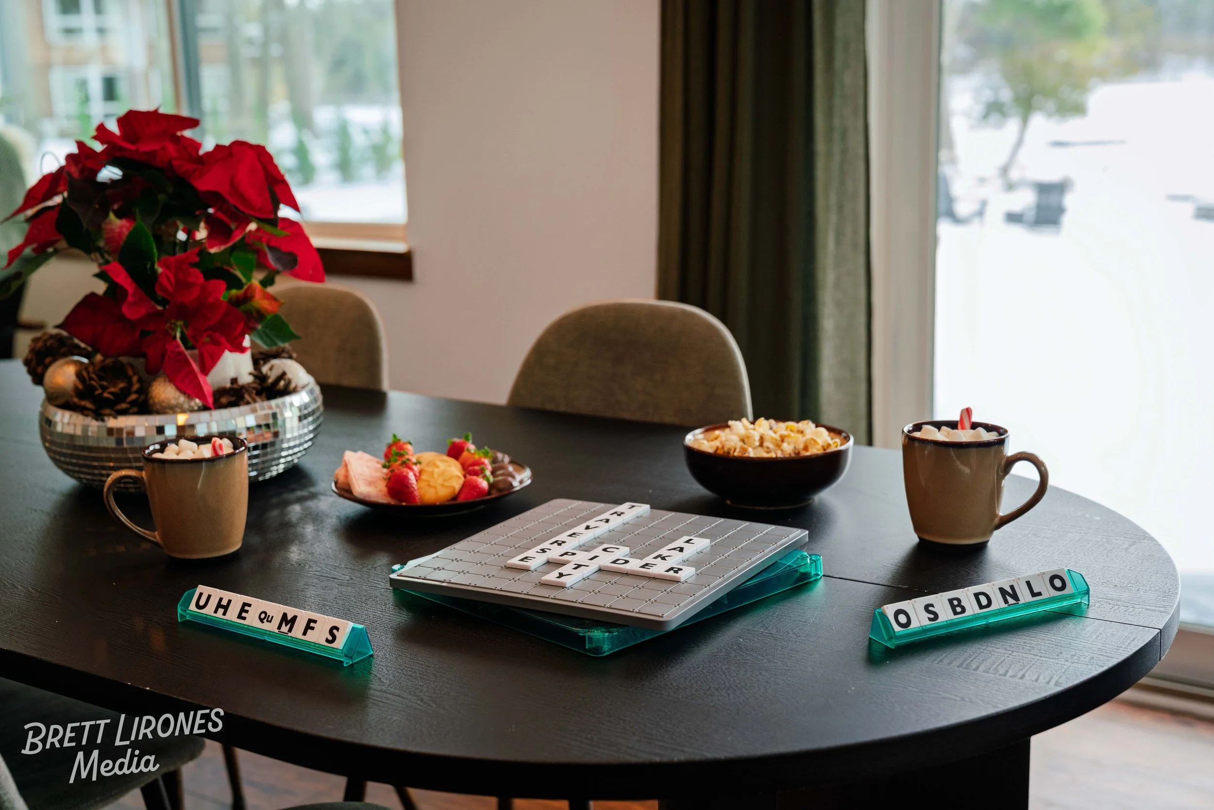 A round black dining table with holiday decorations, including a poinsettia plant, cookies, strawberries with cream, popcorn, and two coffee mugs with marshmallows. Scrabble tiles are arranged on the table and in clear holders, with one row displayin