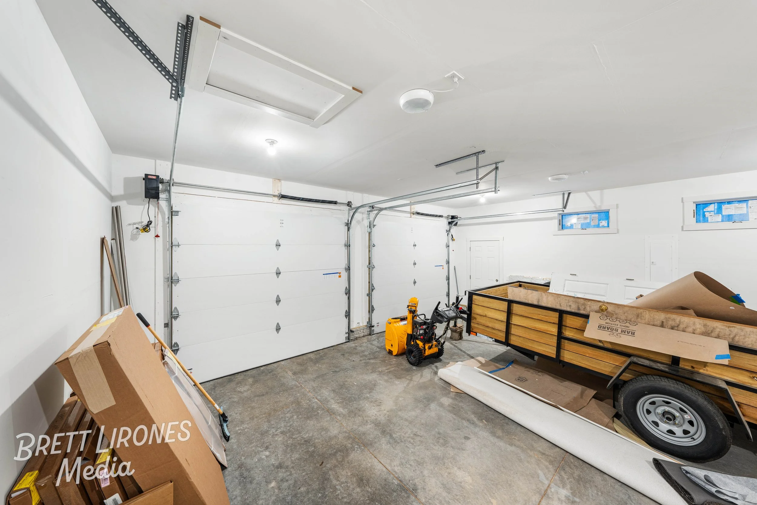 Empty garage with a white door, various boxes and construction materials, a wheelbarrow, and a trailer with plywood boards inside.