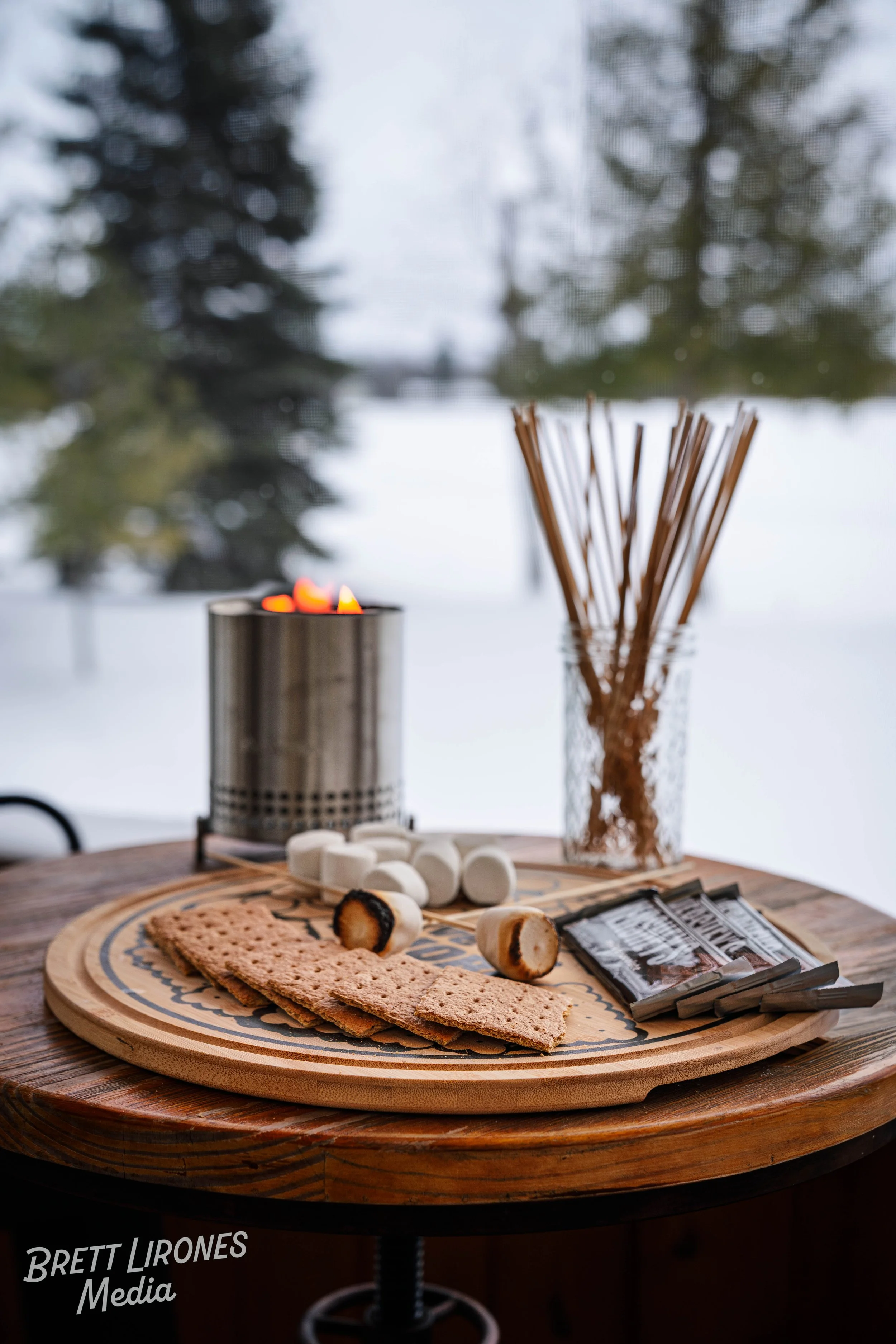 A wooden table with graham crackers, marshmallows, and chocolate for s'mores, set outdoors near a lake or river, with a candle and a jar of wooden sticks in the background, trees, and snow visible.