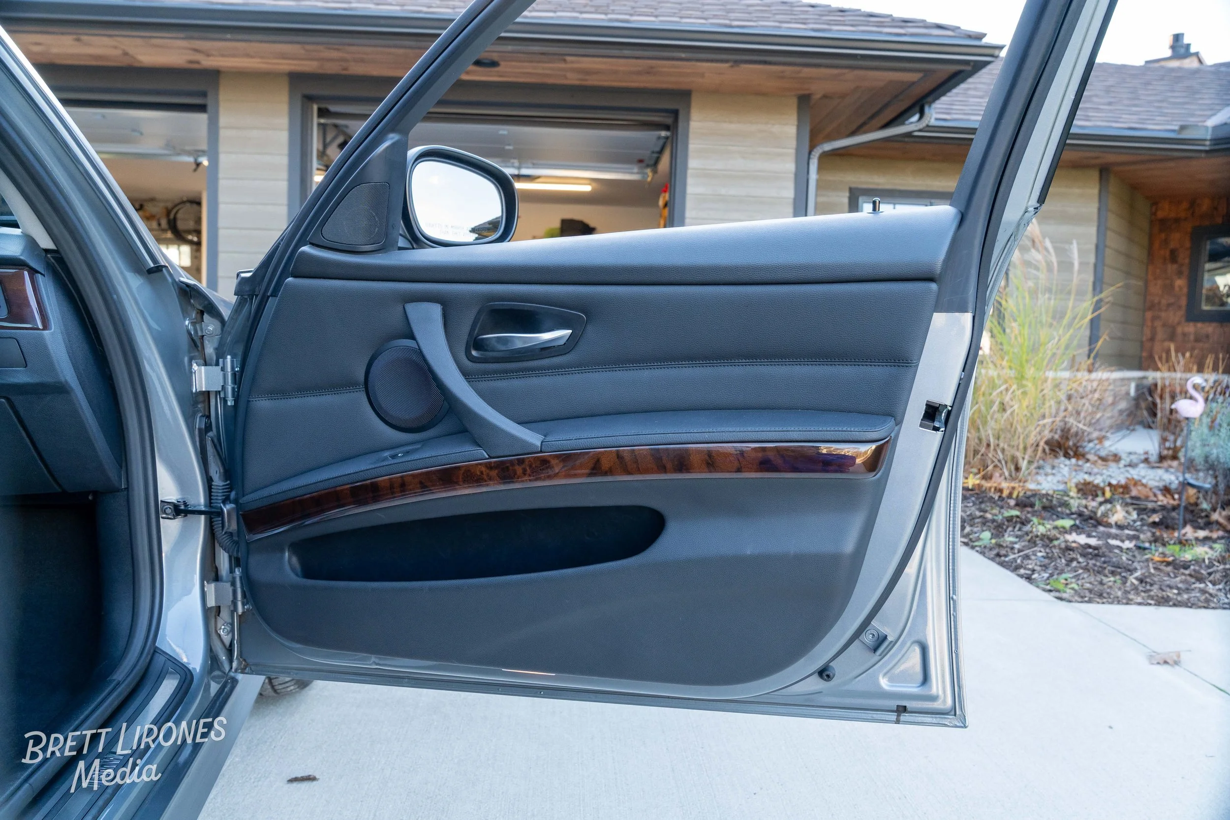The inside of a car door with black interior, a wooden trim, a speaker, window controls, and a door handle, with a house and garden visible outside.
