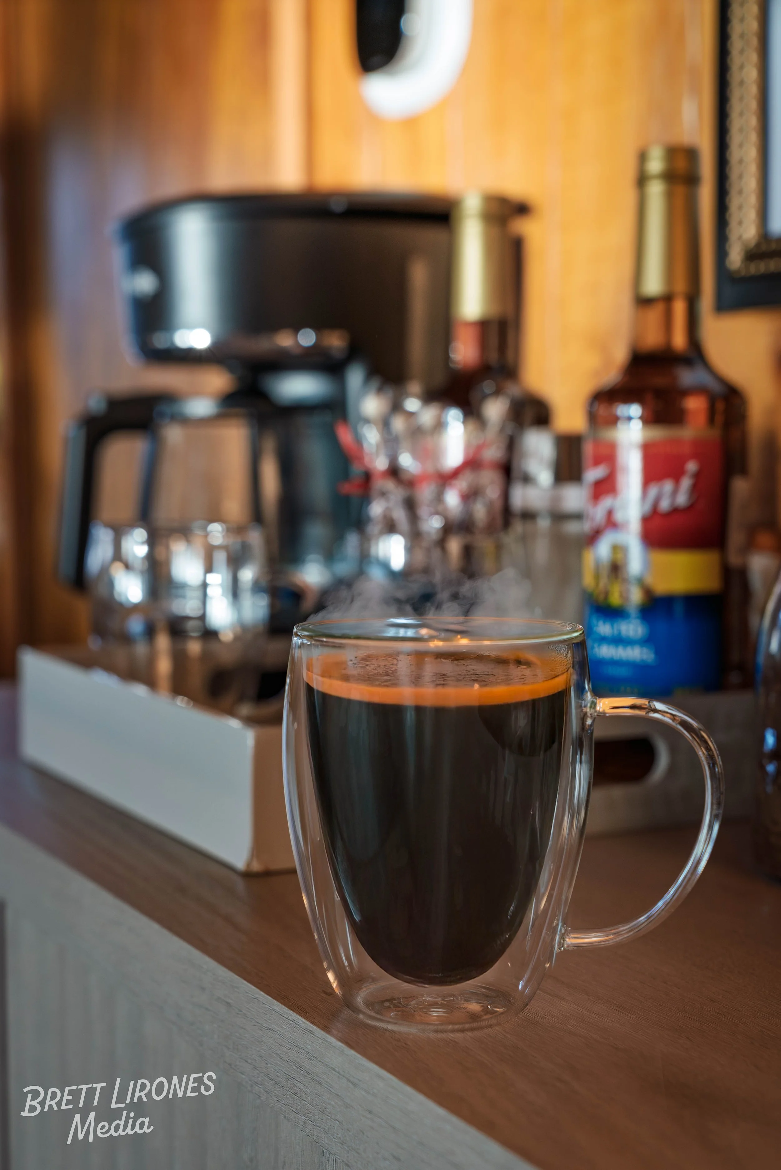 A transparent glass mug filled with hot black coffee on a wooden surface, with a coffee maker and various coffee supplies in the blurred background.