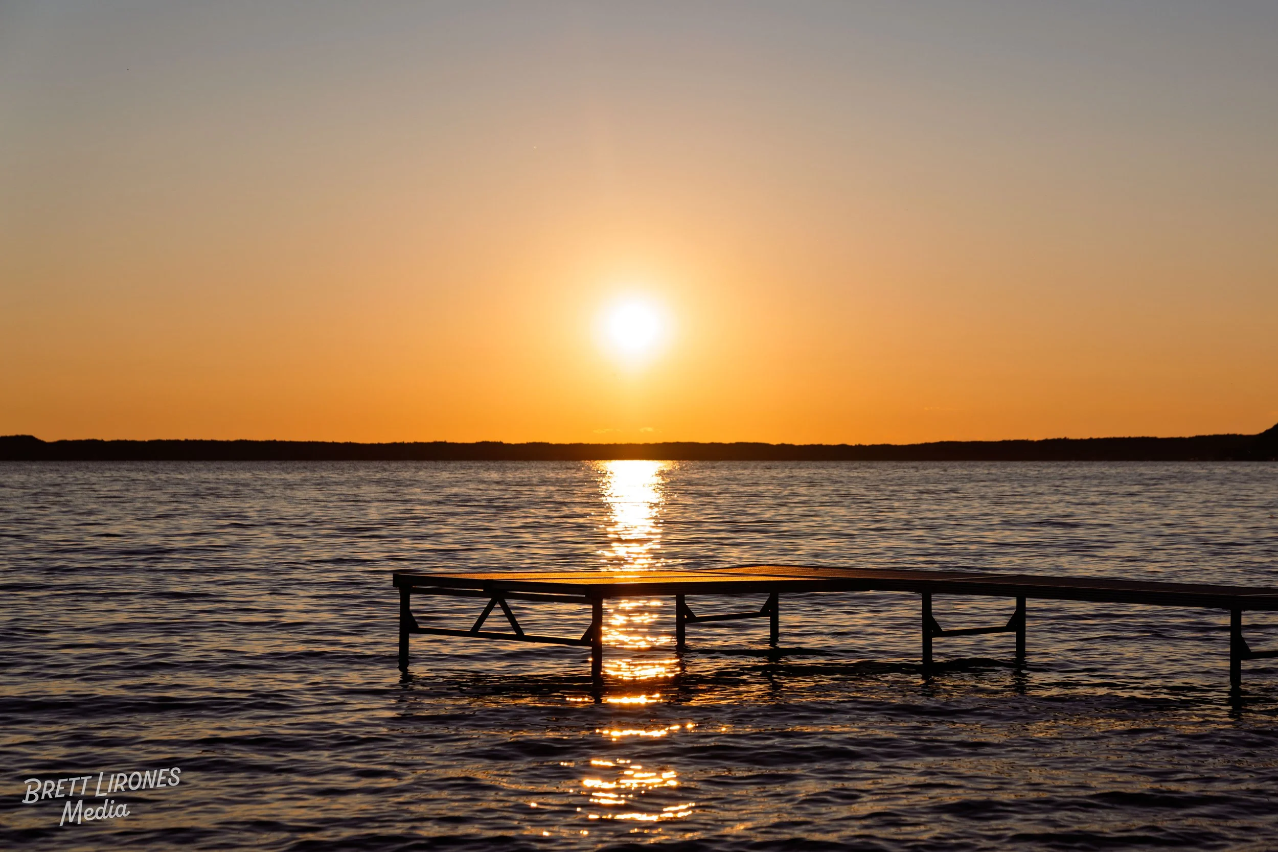 Sunset over water with a partially submerged dock in the foreground and a dark horizon line, orange sky, and reflecting sunlight.