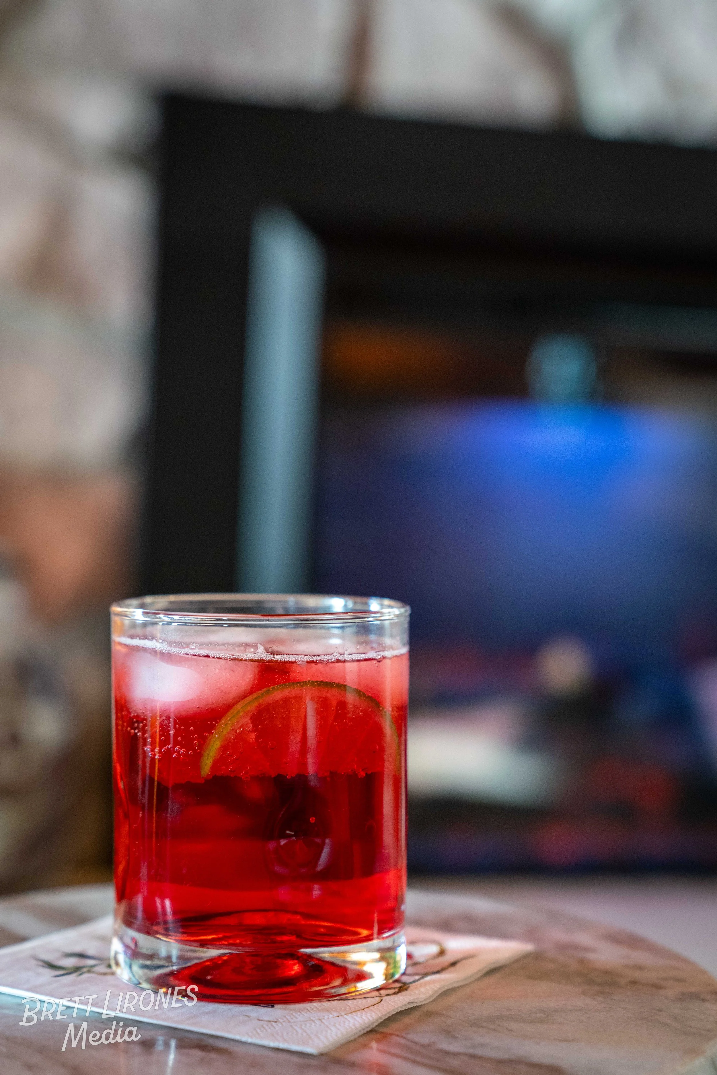 A glass of red beverage with ice and a lemon slice on a napkin on a wooden surface, blurred background with dark and blue tones.