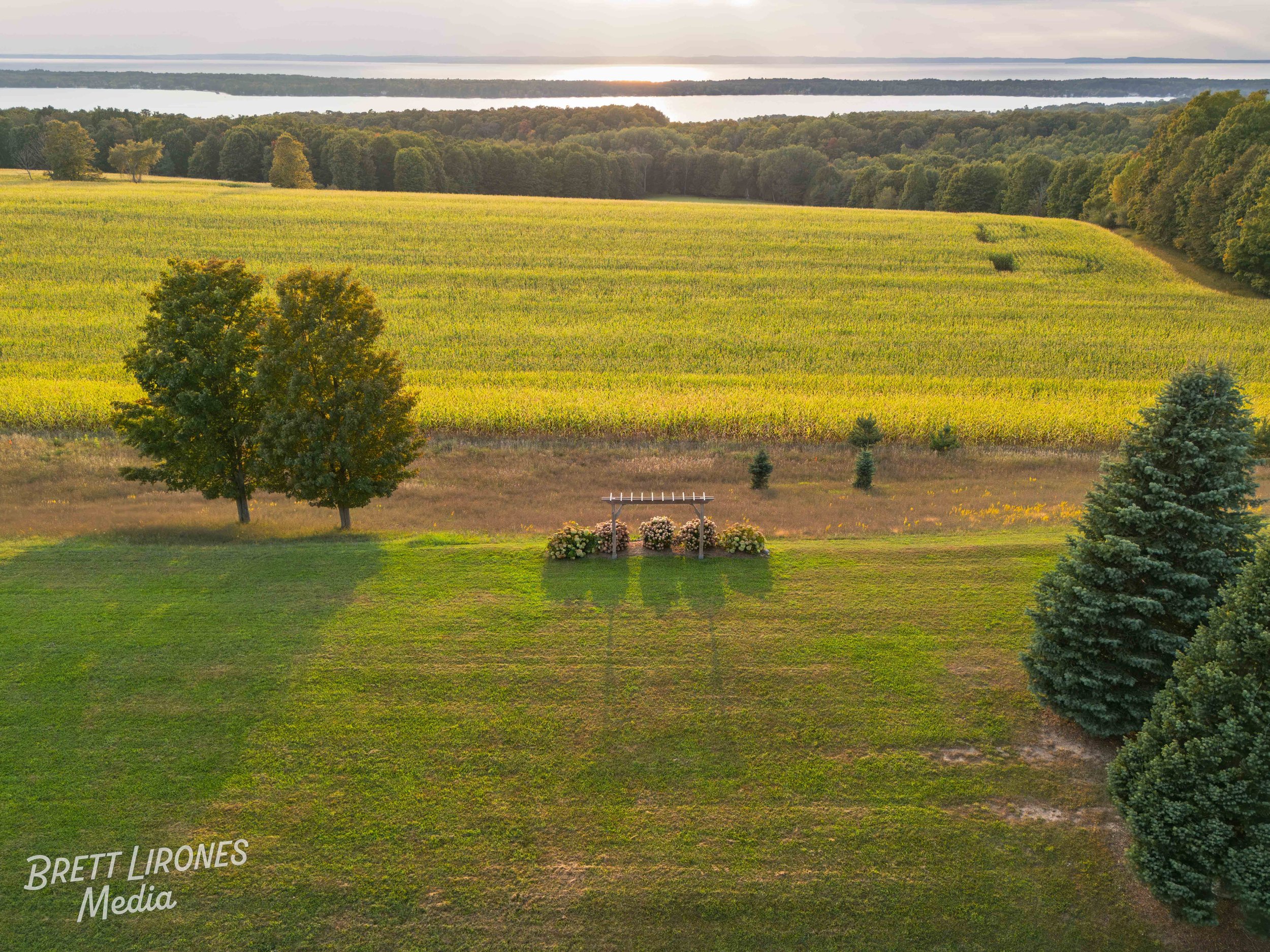 An aerial view of a park with a small monument or plaque surrounded by flowers and trees, with a large field of crops and a river in the background at sunset.