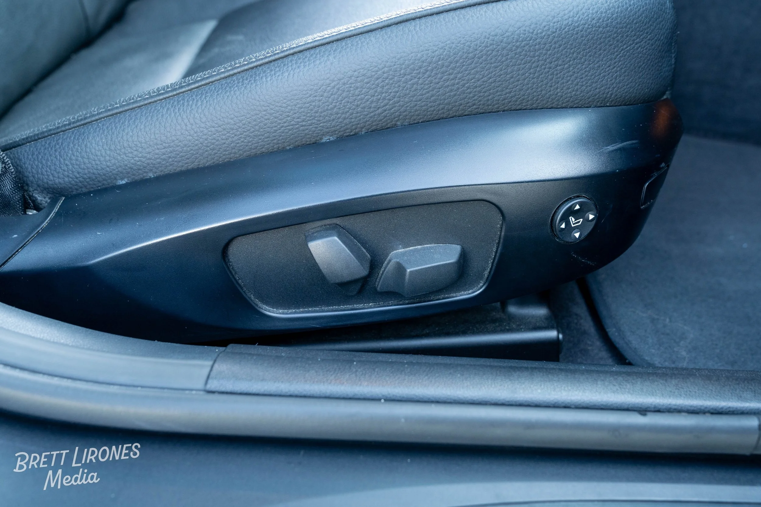 Close-up of a car seat adjustment controls, including two switches and a round button with directional arrows, located on the side of a black leather car seat.