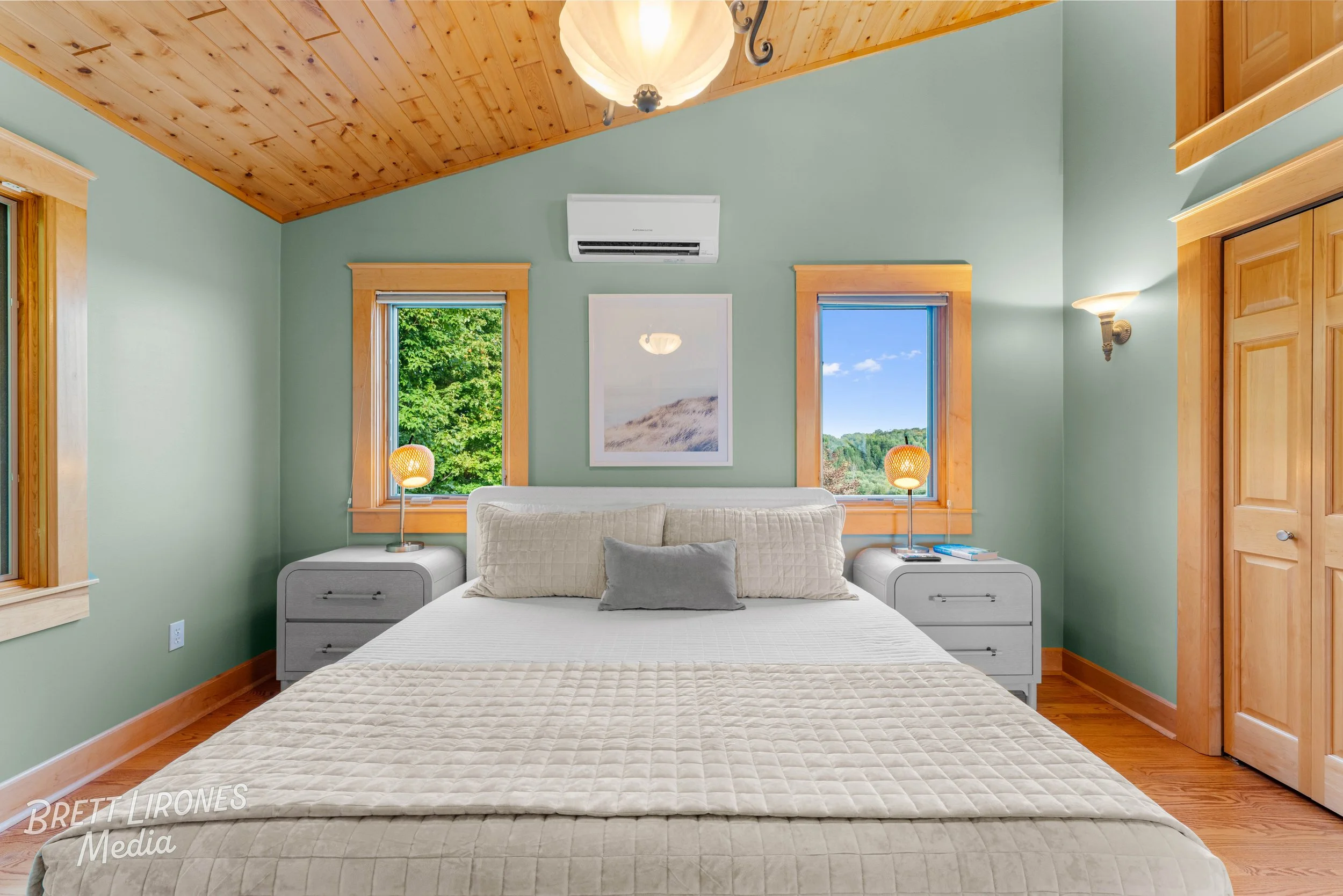 Cozy bedroom with light green walls, wooden ceiling, and hardwood floor. A white bed with a beige quilted blanket and matching pillows is centered in the room. Two nightstands with lamps are on either side of the bed. There are two windows with woode
