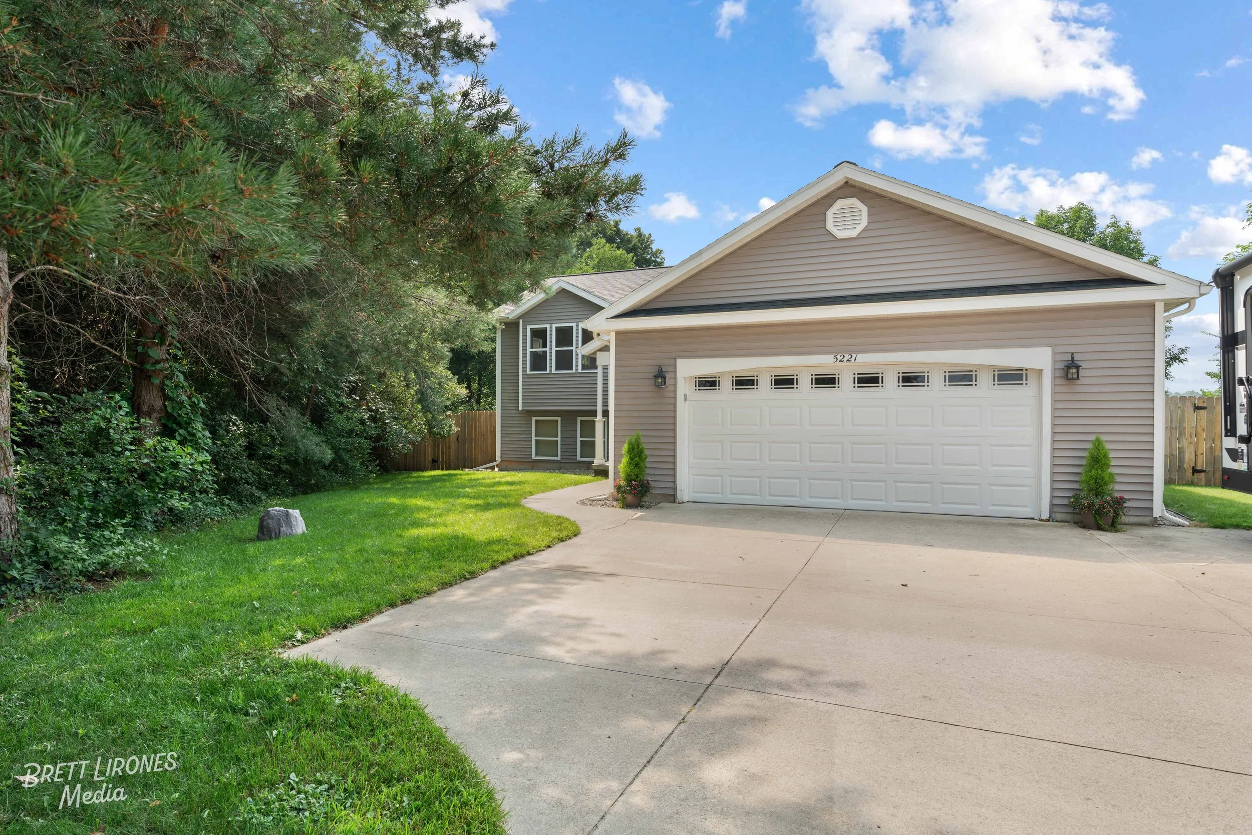 Front of a suburban house with a two-car garage, concrete driveway, and green lawn, surrounded by trees and shrubs, under a blue sky with some clouds.
