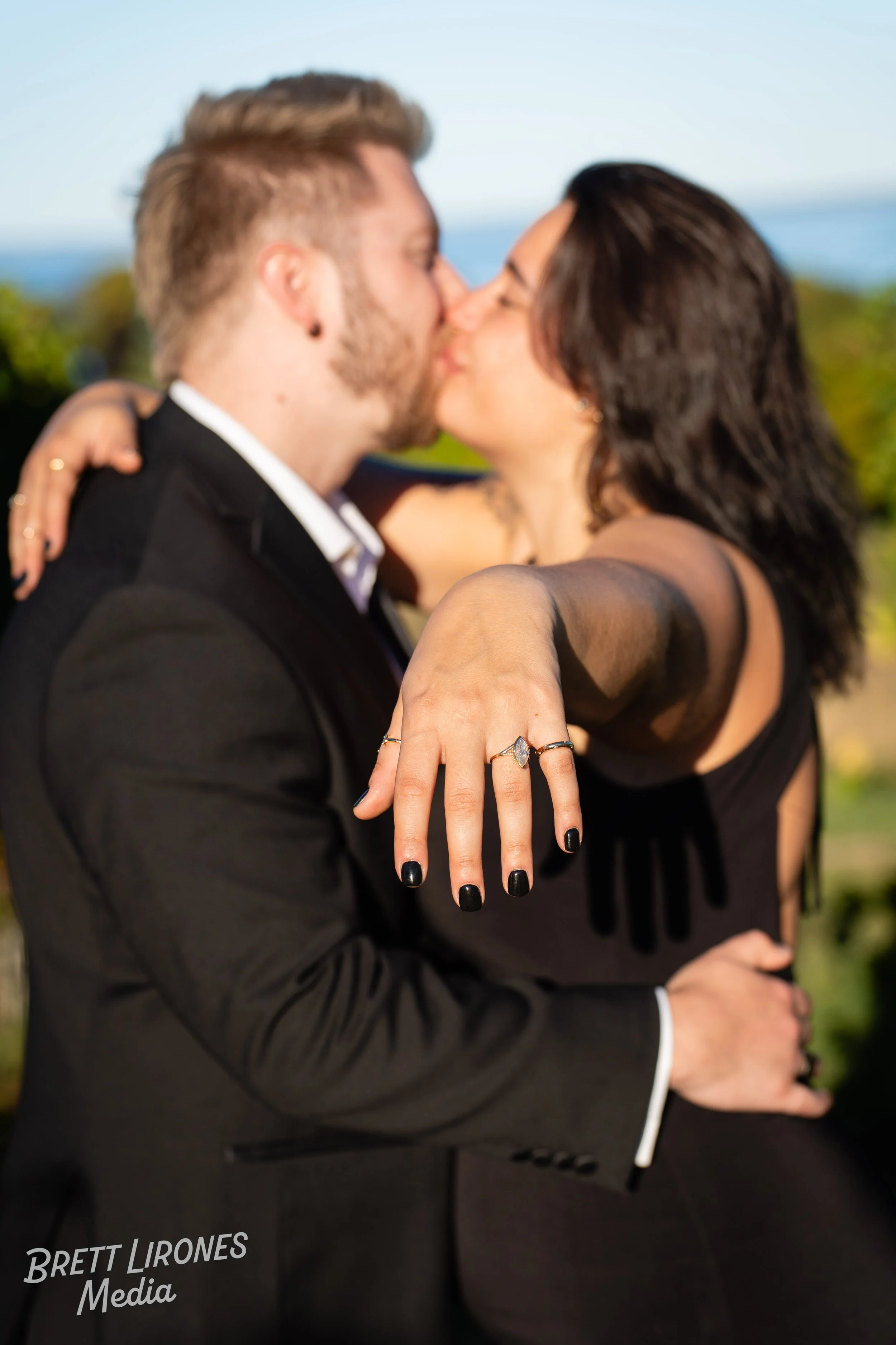 A couple in formal attire sharing a kiss outdoors with the woman showing off her engagement ring.