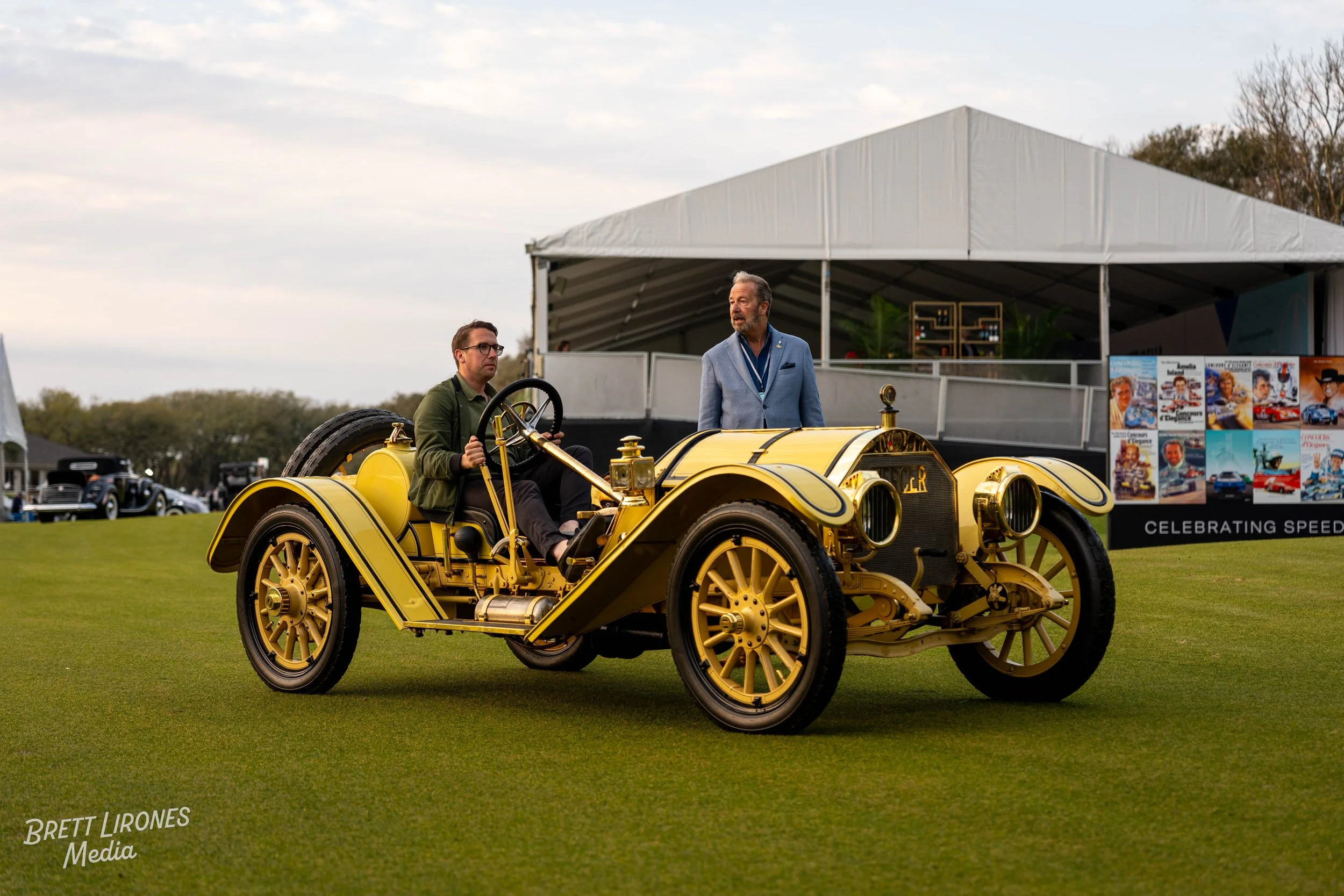 A vintage yellow car with a man in green jacket sitting at the wheel and another man in blue blazer standing nearby at an outdoor event.