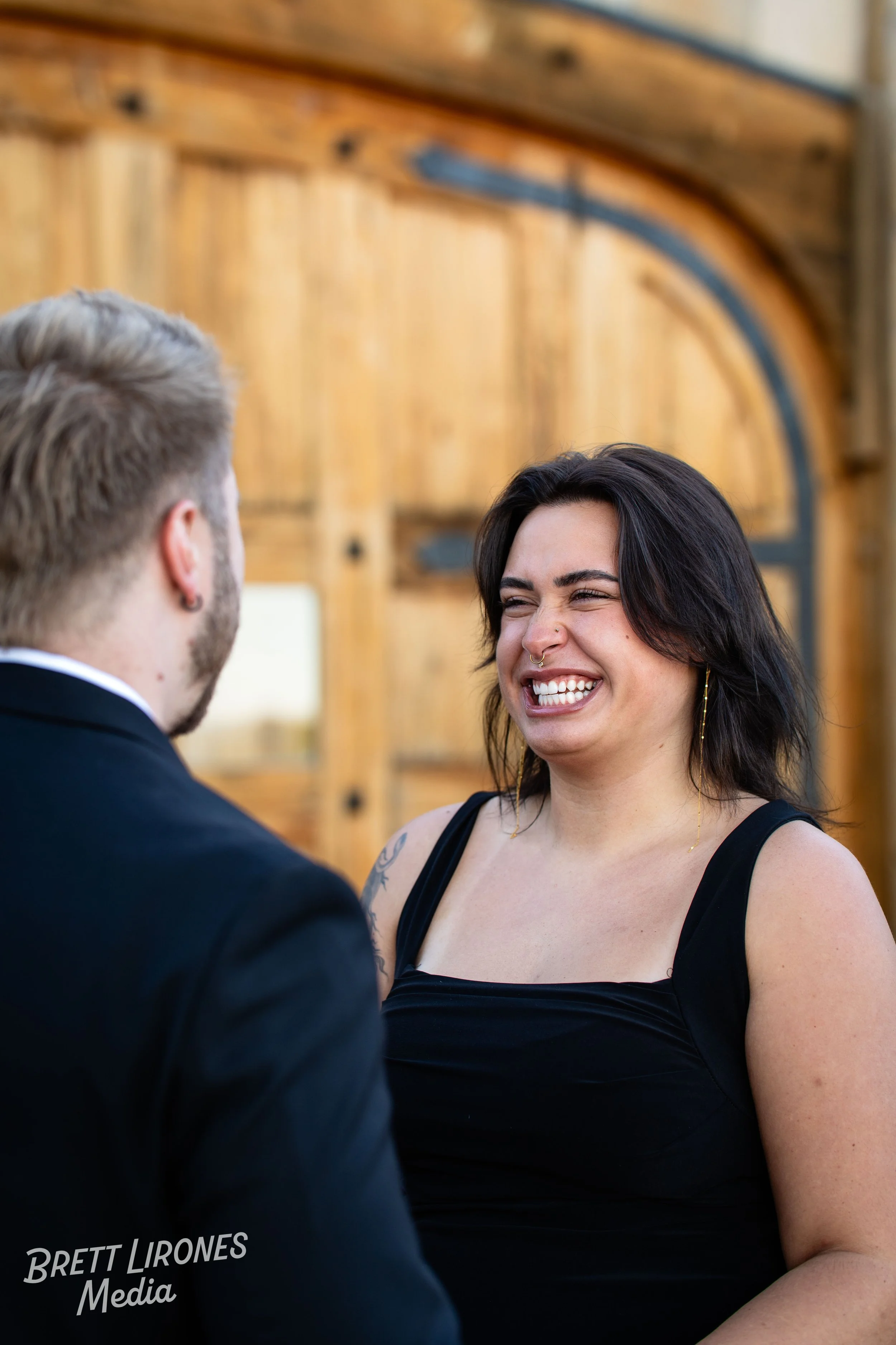 A woman with dark hair, wearing a black top, is smiling and facing a man in a suit in front of a wooden barn door.