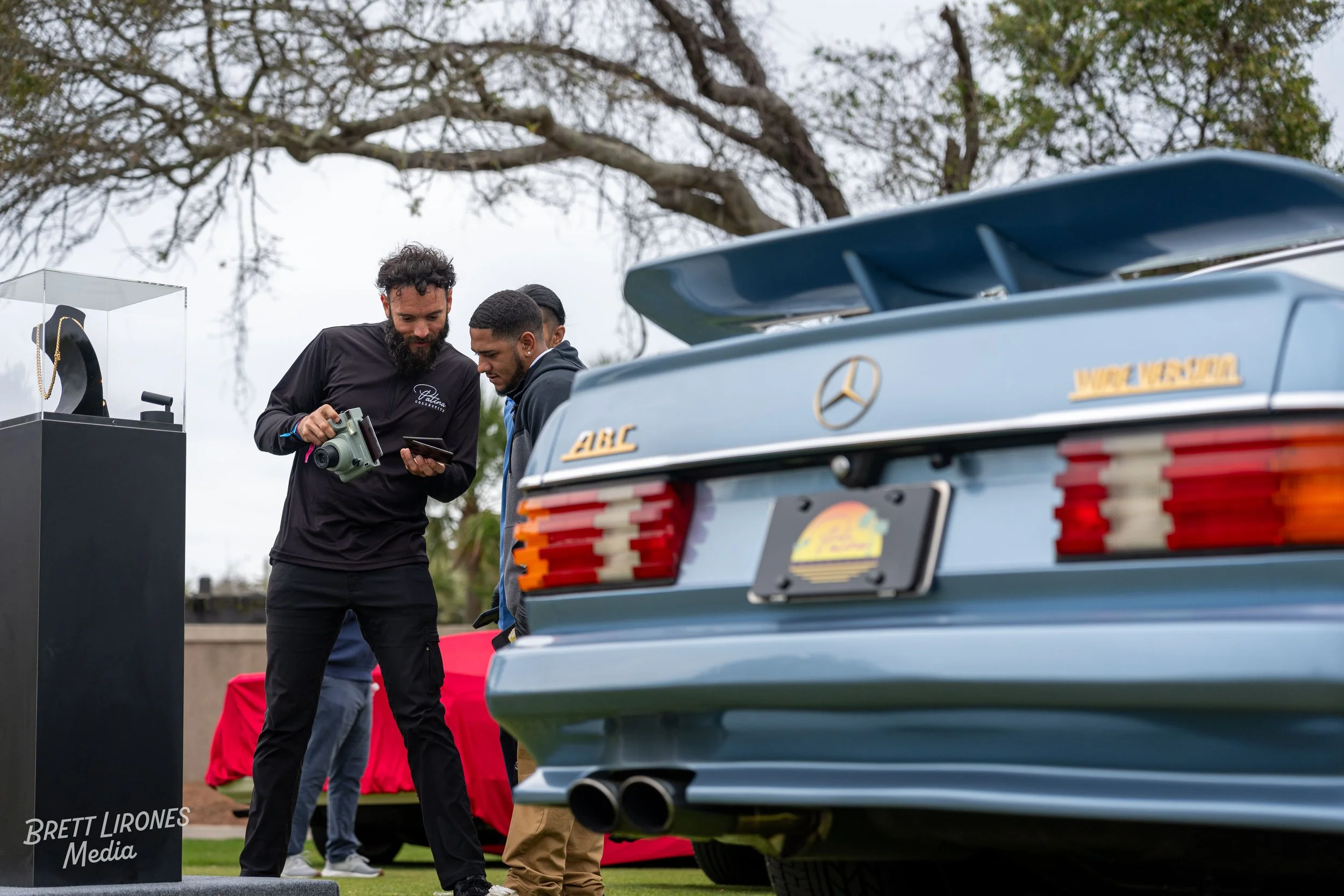 Three men looking at a device or phone near a vintage Mercedes-Benz 300SL car at an outdoor event.