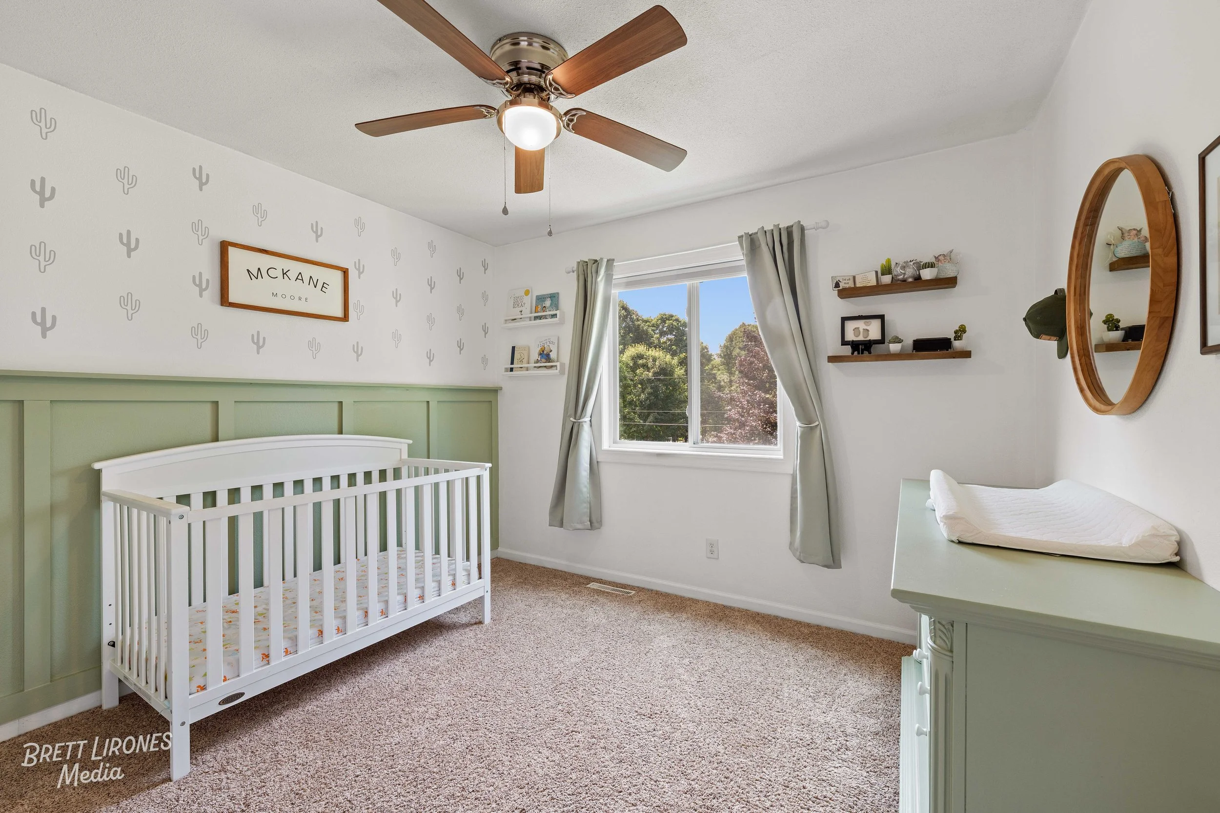 A baby's nursery with a green and white crib, beige carpet, a window with white curtains, and walls decorated with small gray cactus patterns and shelves holding books and decor. There is a ceiling fan with wooden blades and a light, and a changing t