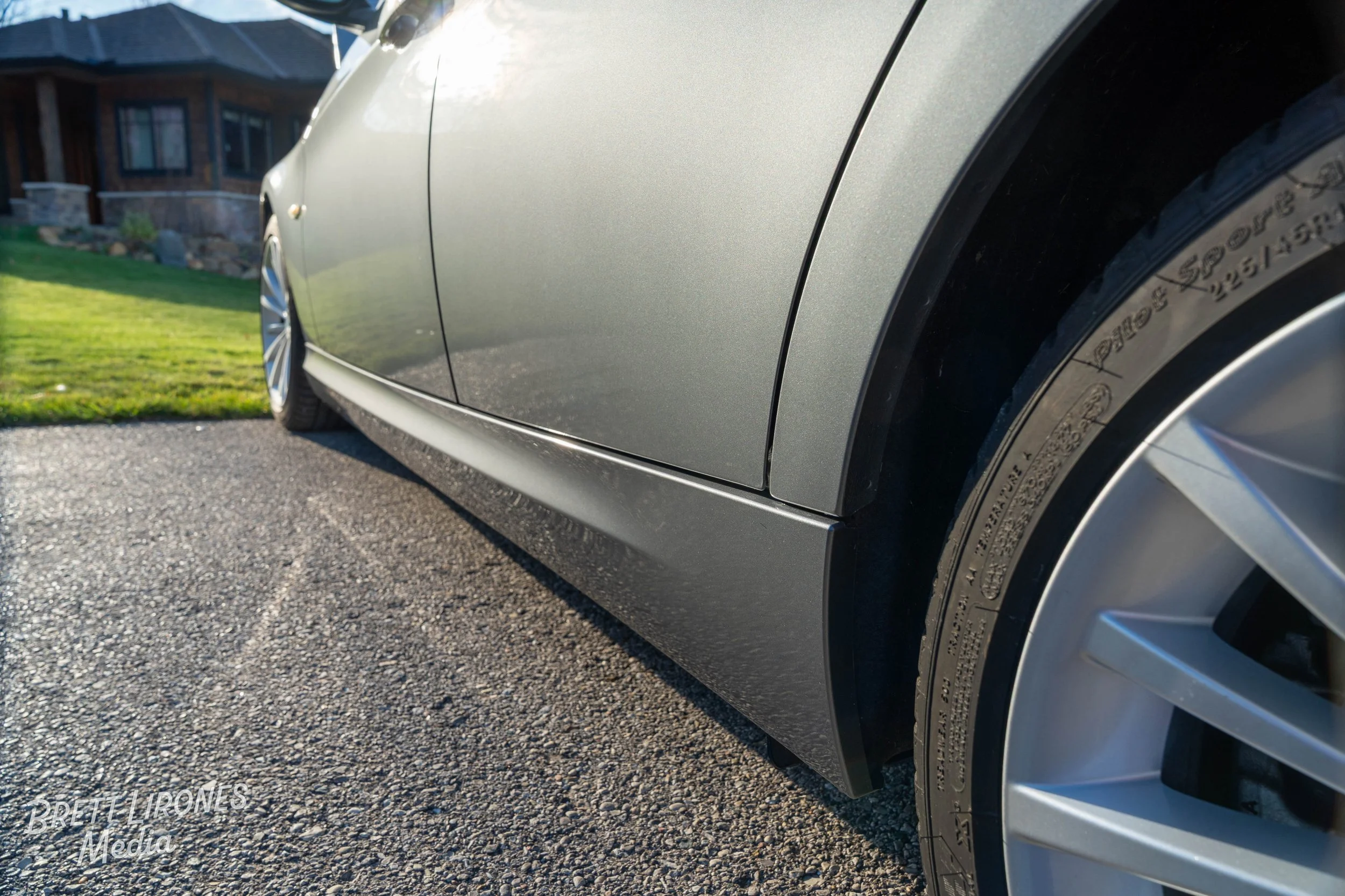 Close-up of a gray car's side, focusing on the front wheel and body, parked on a driveway with a house and greenery in the background.