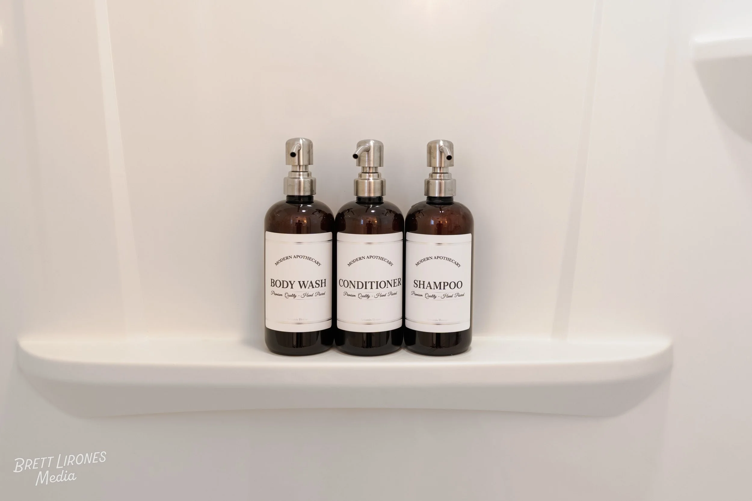 Three travel-sized bottles of toiletries labeled as body wash, conditioner, and shampoo on a white shower shelf.