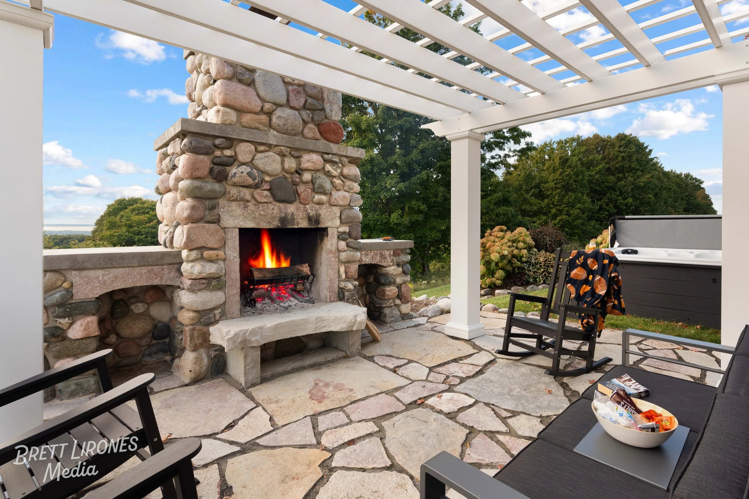 A backyard patio with a stone fireplace with a fire burning, a hot tub, a black rocking chair with a blanket, a gray sofa with snacks, and a white pergola overhead, green trees in the background.