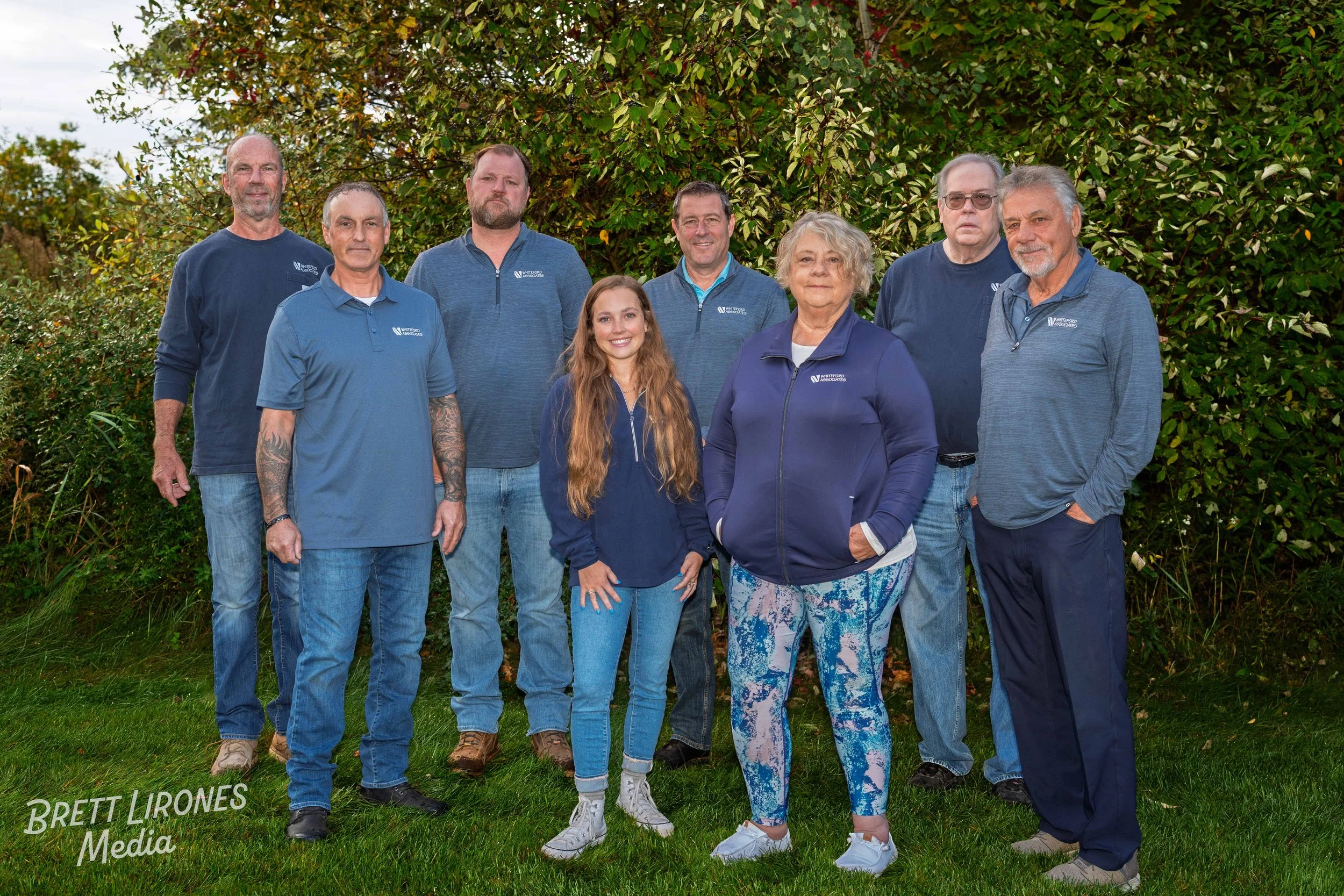 Group of nine people standing outdoors on grass in front of a bush, dressed in blue casual clothing with the 'Anthem Associated' logo, posing for a group photo.