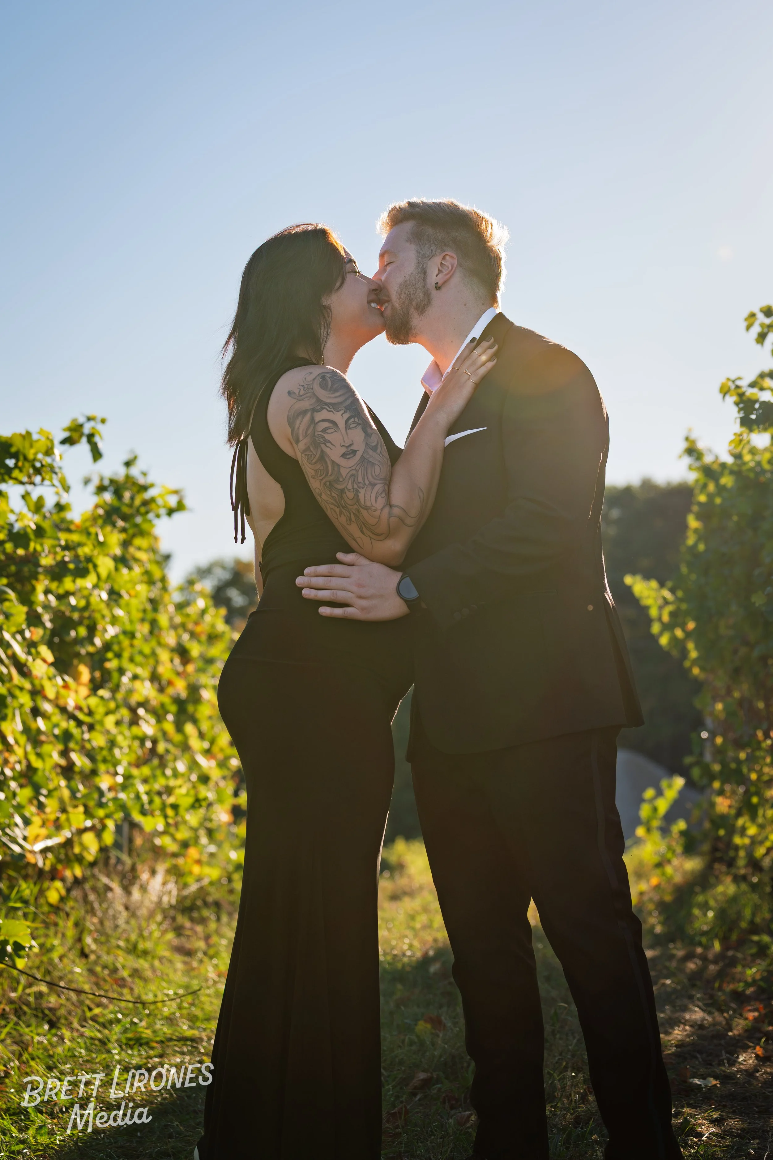 A couple in formal attire kissing outdoors on a sunny day with greenery and a clear blue sky.