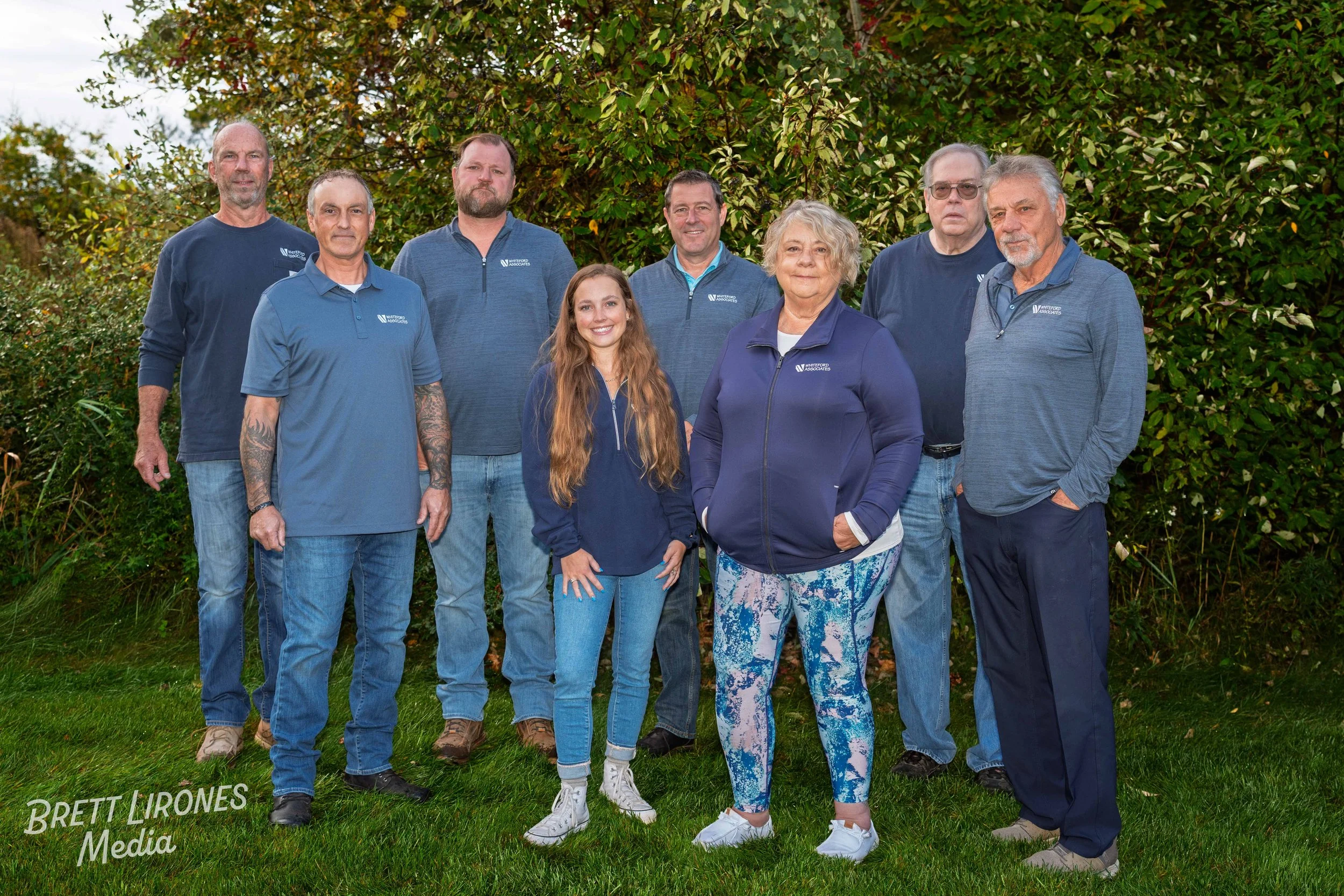 A group of nine people standing outdoors on grass with trees and bushes behind them, wearing casual clothing and smiling.