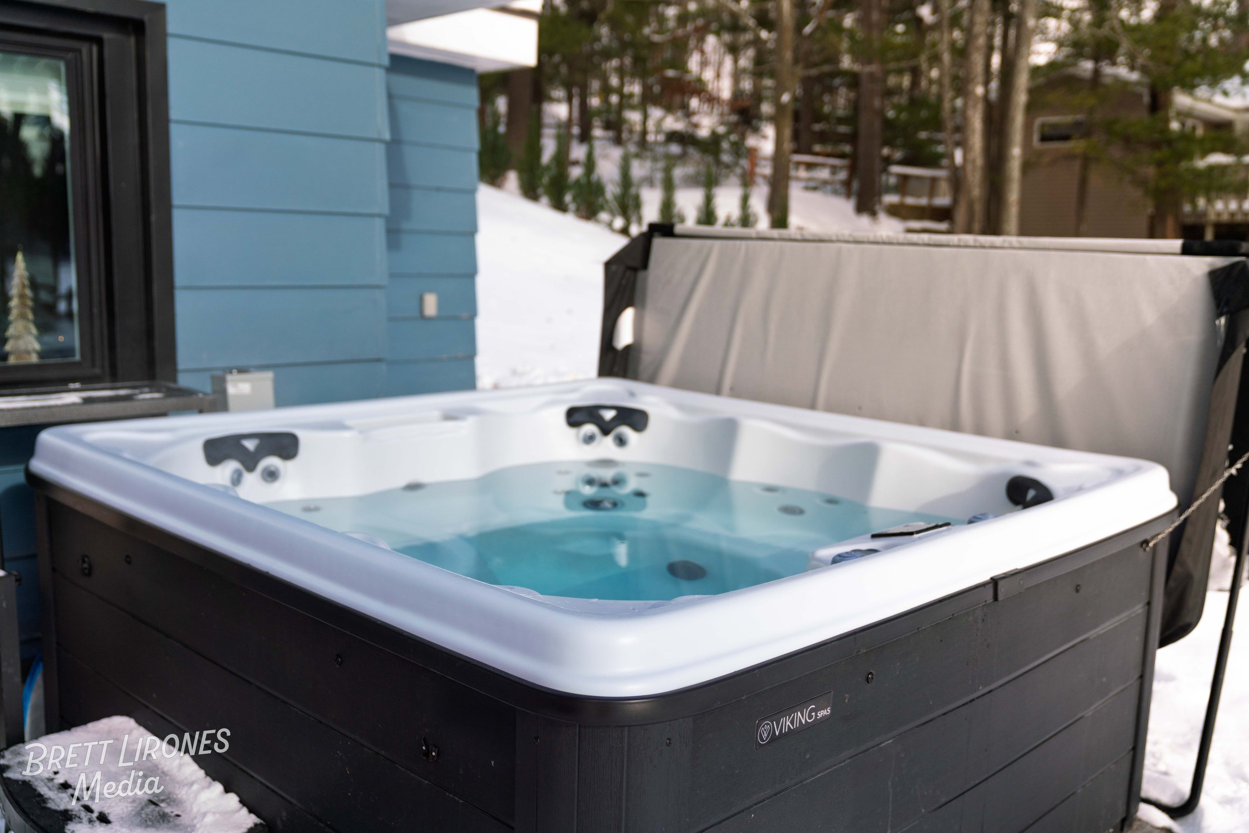 An outdoor hot tub on a snowy deck beside a blue house, surrounded by trees with a covered seating area nearby.