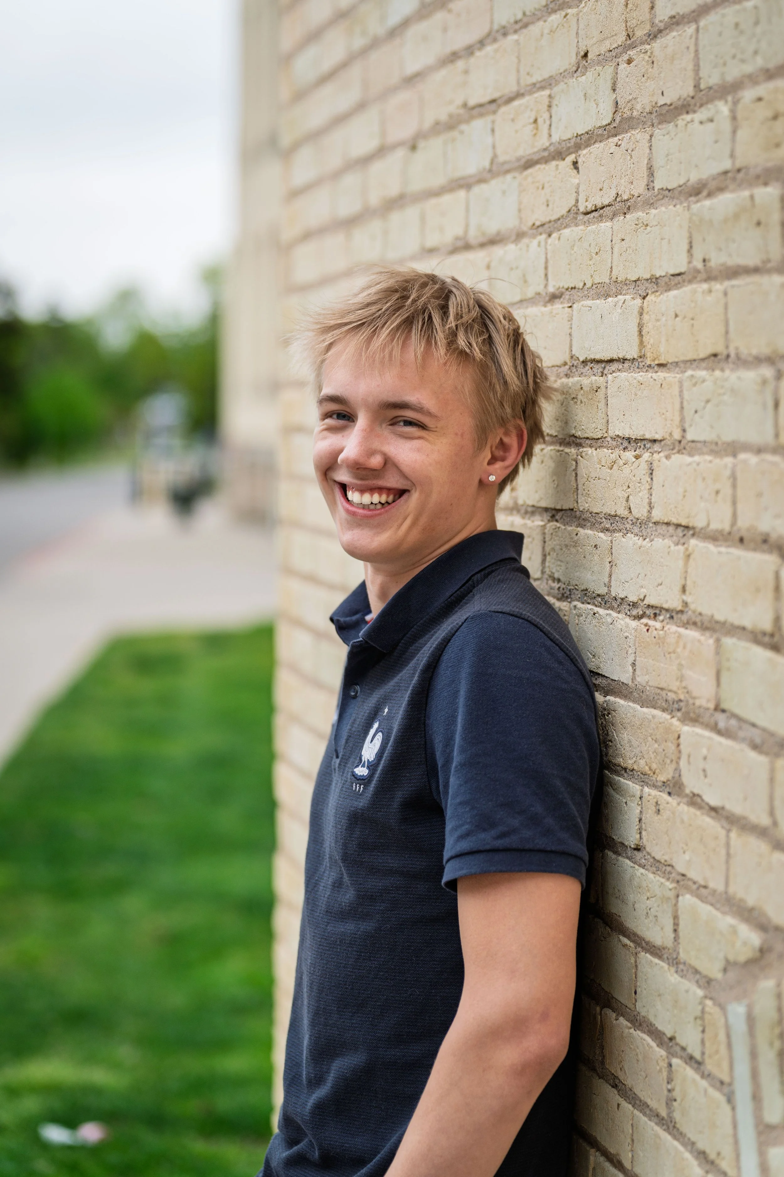Young man with blond hair leaning against a brick wall, smiling, wearing a navy blue polo shirt, outdoors during daytime.