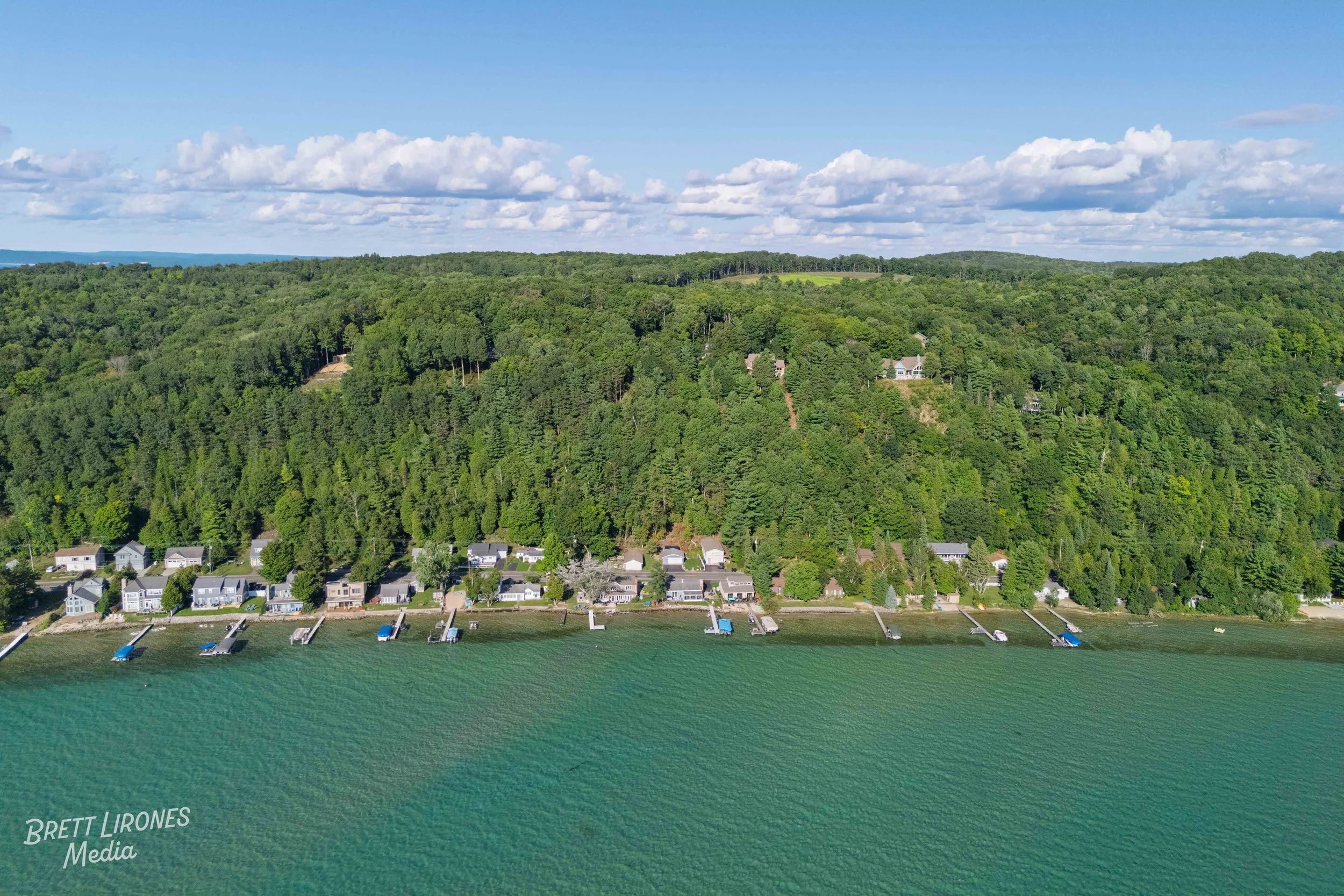 Aerial view of a lakeside community with houses along the shoreline, private docks extending into the water, and a densely wooded green hill in the background under a partly cloudy sky.