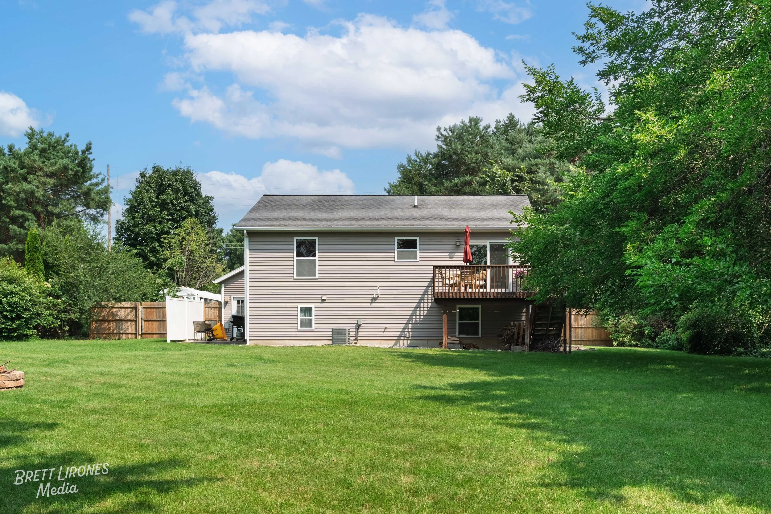 Backyard view of a two-story house with vinyl siding, a small deck with a table and umbrella, surrounded by trees and a wooden fence, under a partly cloudy sky.