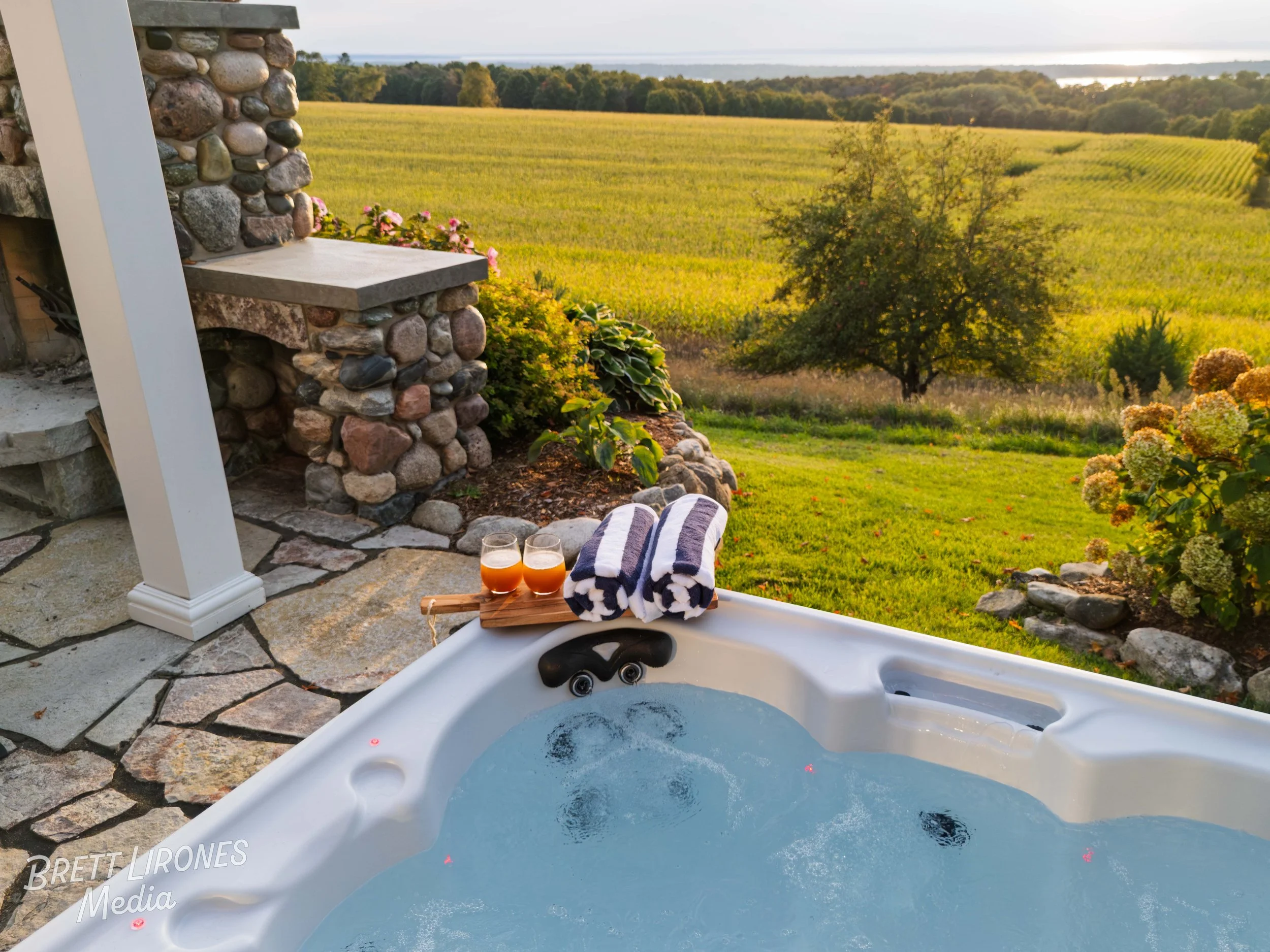 A hot tub with bubbling water on a patio overlooking a grassy field and trees during sunset. On the edge of the hot tub, there are two glasses of orange beverage, two rolled towels, and a small wooden tray.
