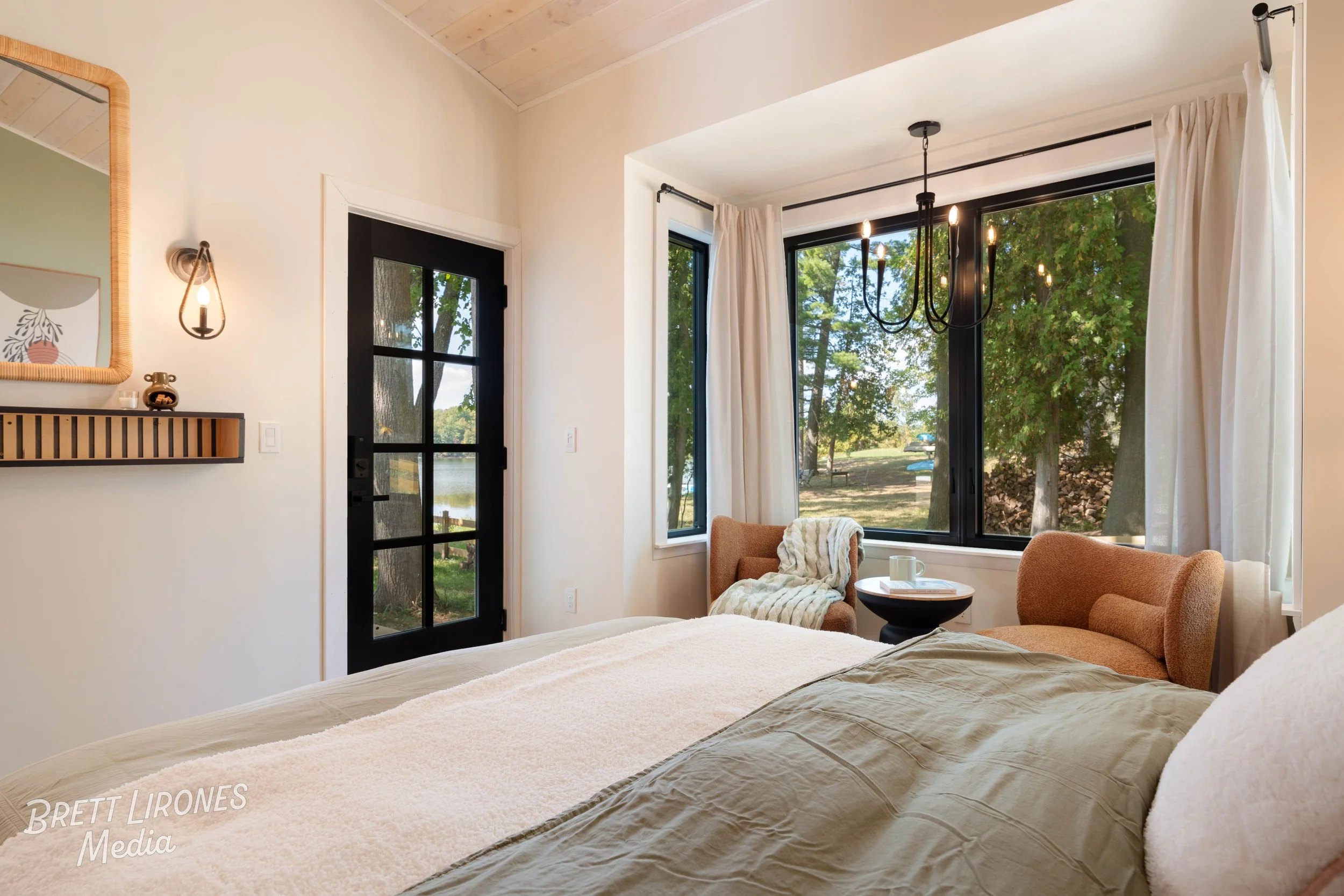 A bedroom with large windows showing a view of trees and a lake outside. The room has a bed with beige and white linens, a black door with glass panes, a black chandelier hanging from the ceiling, and cozy seating including a chair with a blanket, an