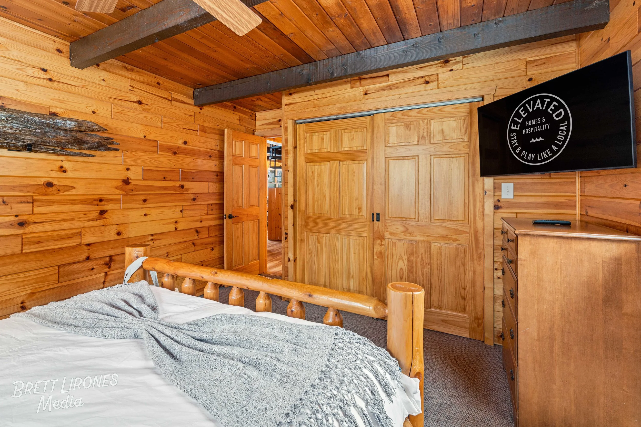 A rustic bedroom with wooden walls, ceiling, and furniture, featuring a bed with a gray blanket, a closed wooden door, a wall-mounted TV, and a dresser.