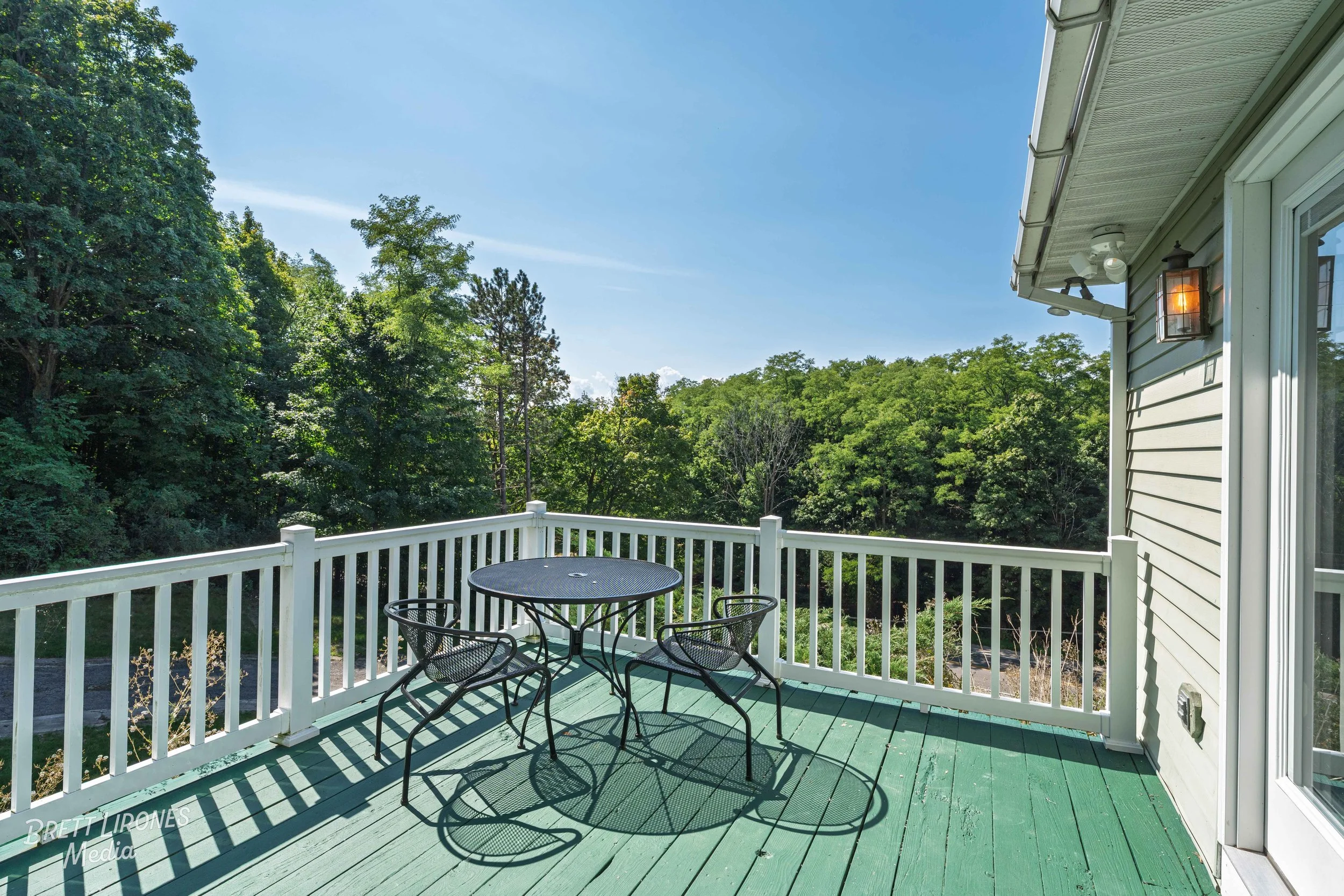 A wooden outdoor balcony with a black metal table and four matching chairs overlooking a lush green forest under a blue sky.