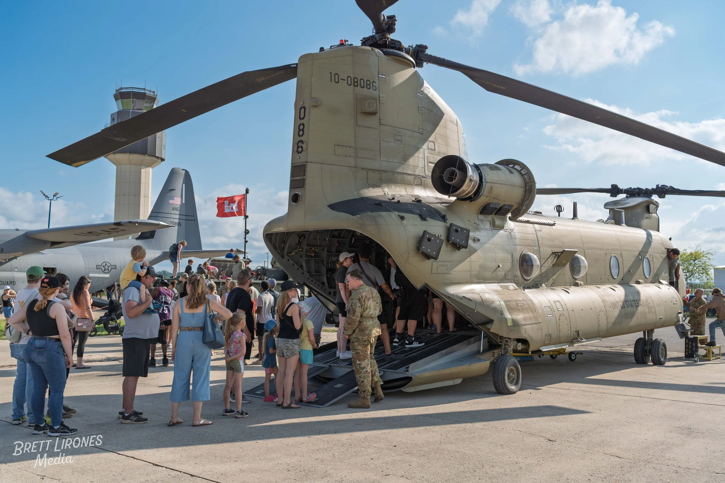 Children and adults visiting a military helicopter exhibit outdoors on a sunny day. The helicopter is beige with an open cargo door, and there is a crowd around it. Other aircraft and control tower are visible in the background.